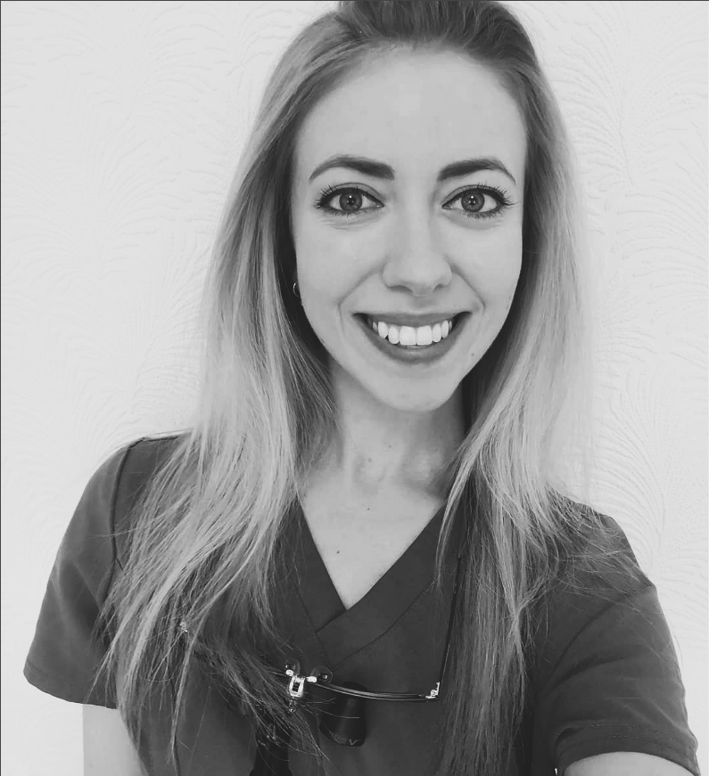 Black and white photo of a smiling woman with long hair wearing scrubs and glasses hanging on her neck, standing against a textured wall.