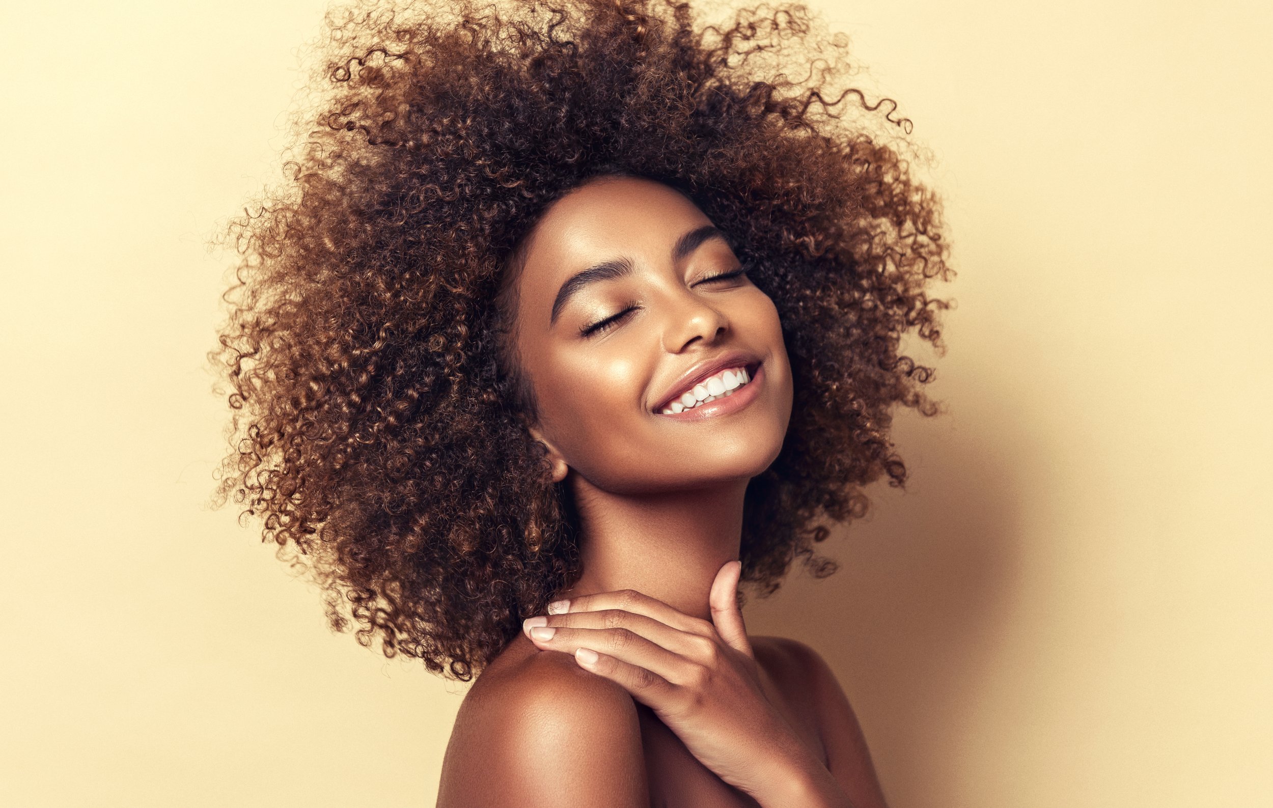 Smiling woman with curly hair, eyes closed, touching her shoulder with one hand against a beige background.