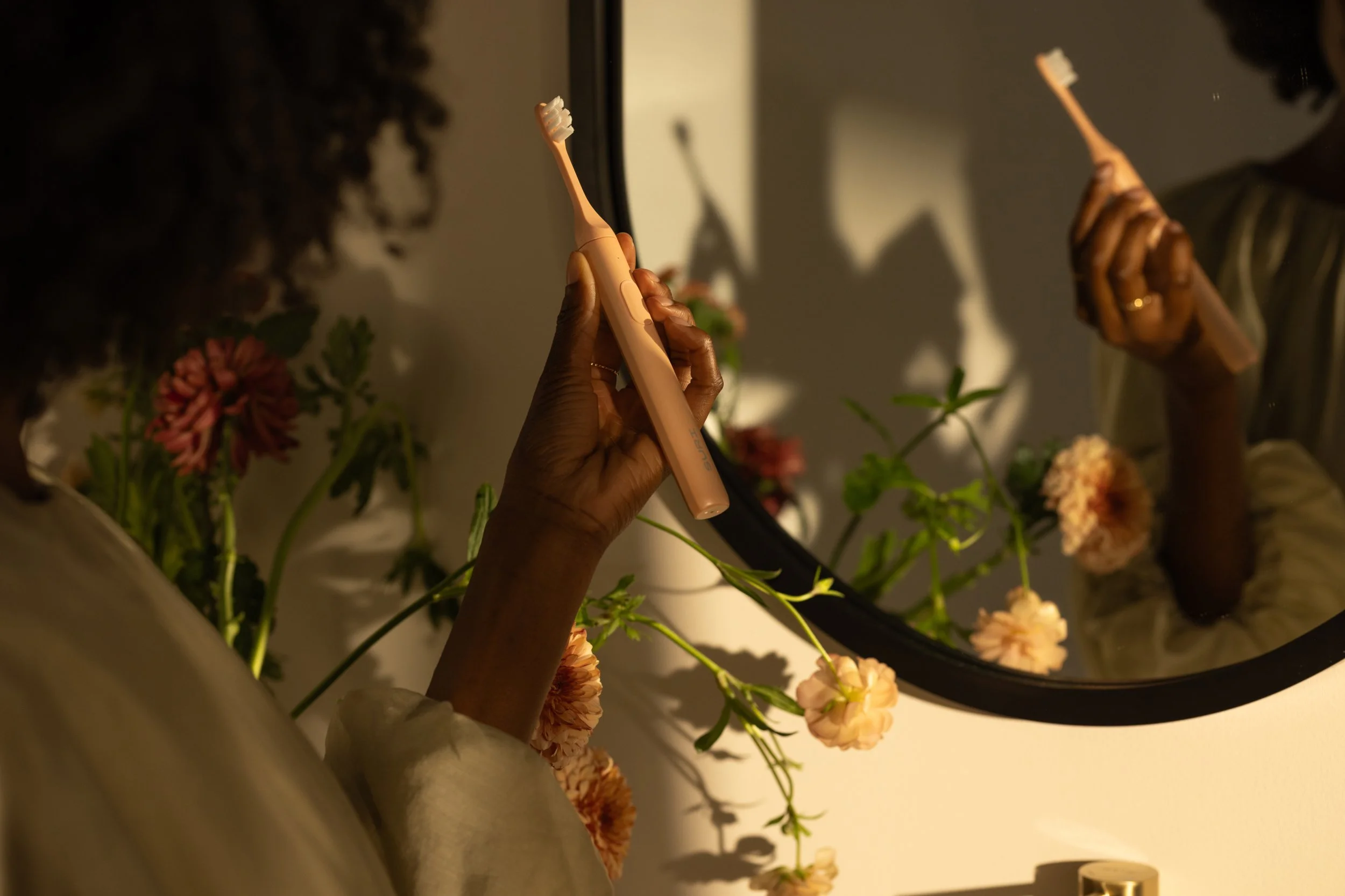 Person brushing teeth with electric toothbrush in front of a mirror, with flower decoration around the mirror.