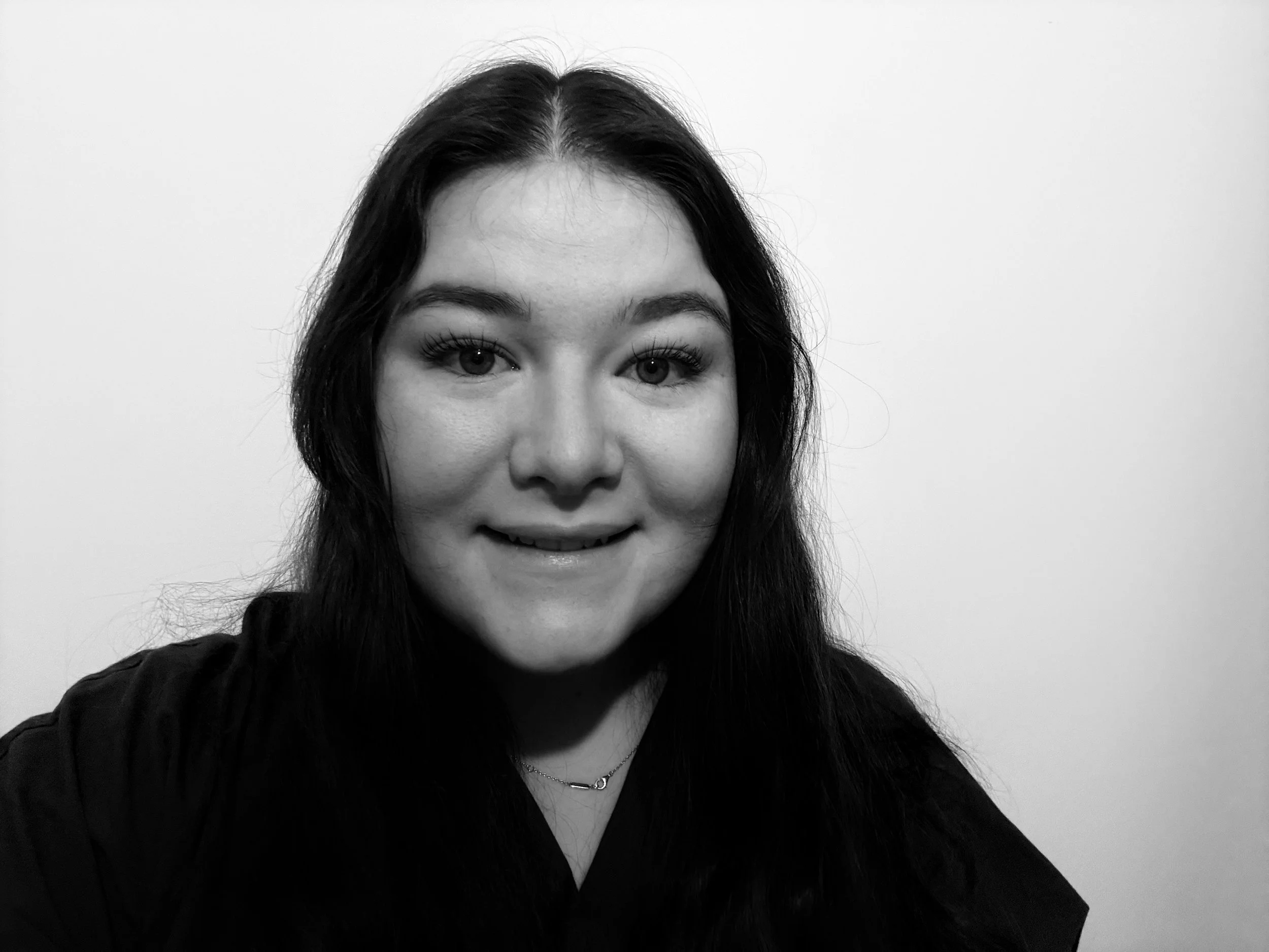 Black and white portrait of a young woman with long dark hair, smiling slightly, wearing a dark top and a delicate necklace, in front of a plain light-colored wall.