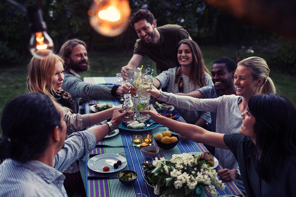 Group of friends having a dinner party outdoors in the evening, raising glasses for a toast, surrounded by food, drinks, and candles on the table.