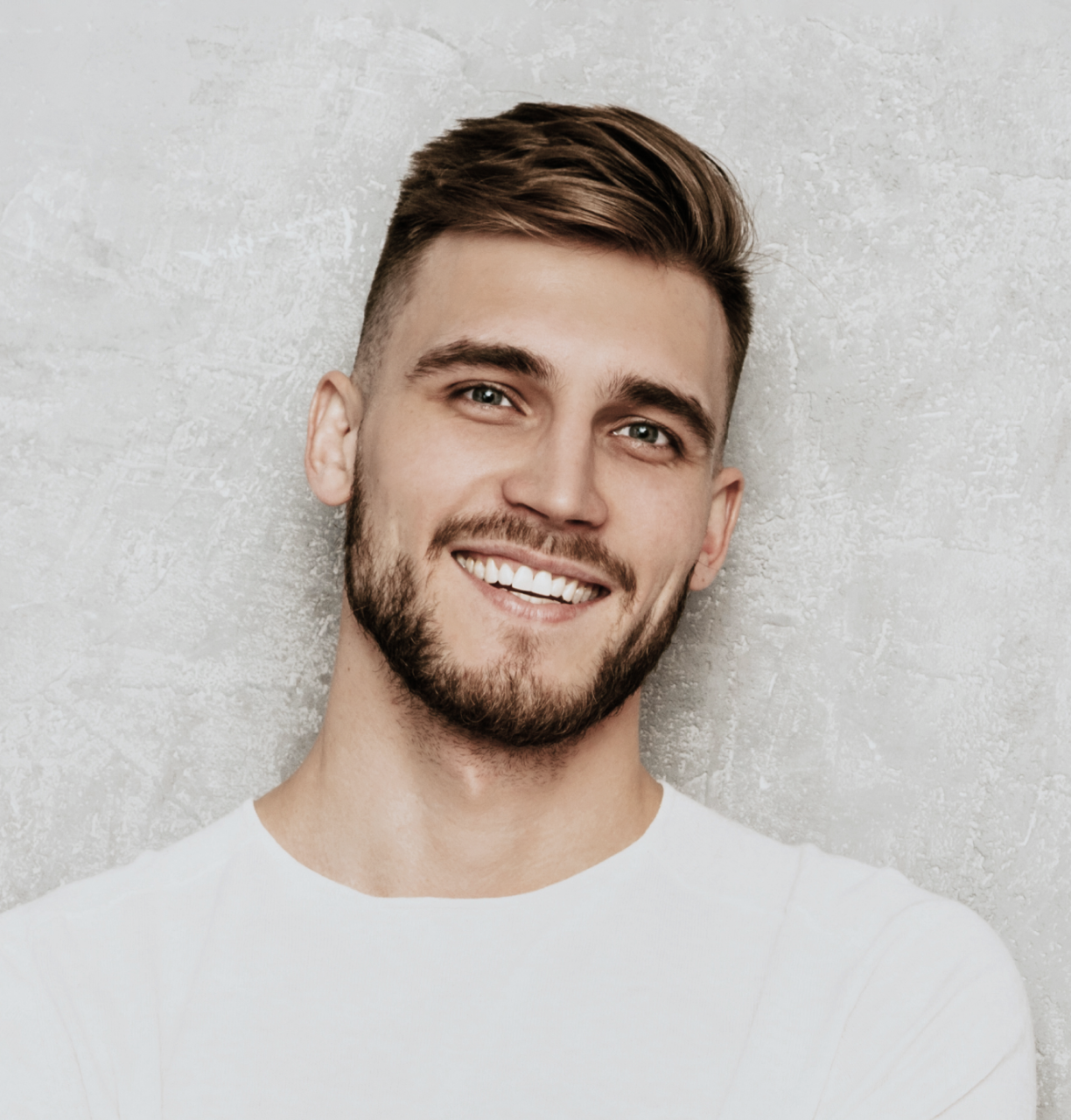 A young man with short brown hair and a beard smiling, wearing a white shirt against a textured gray wall background.