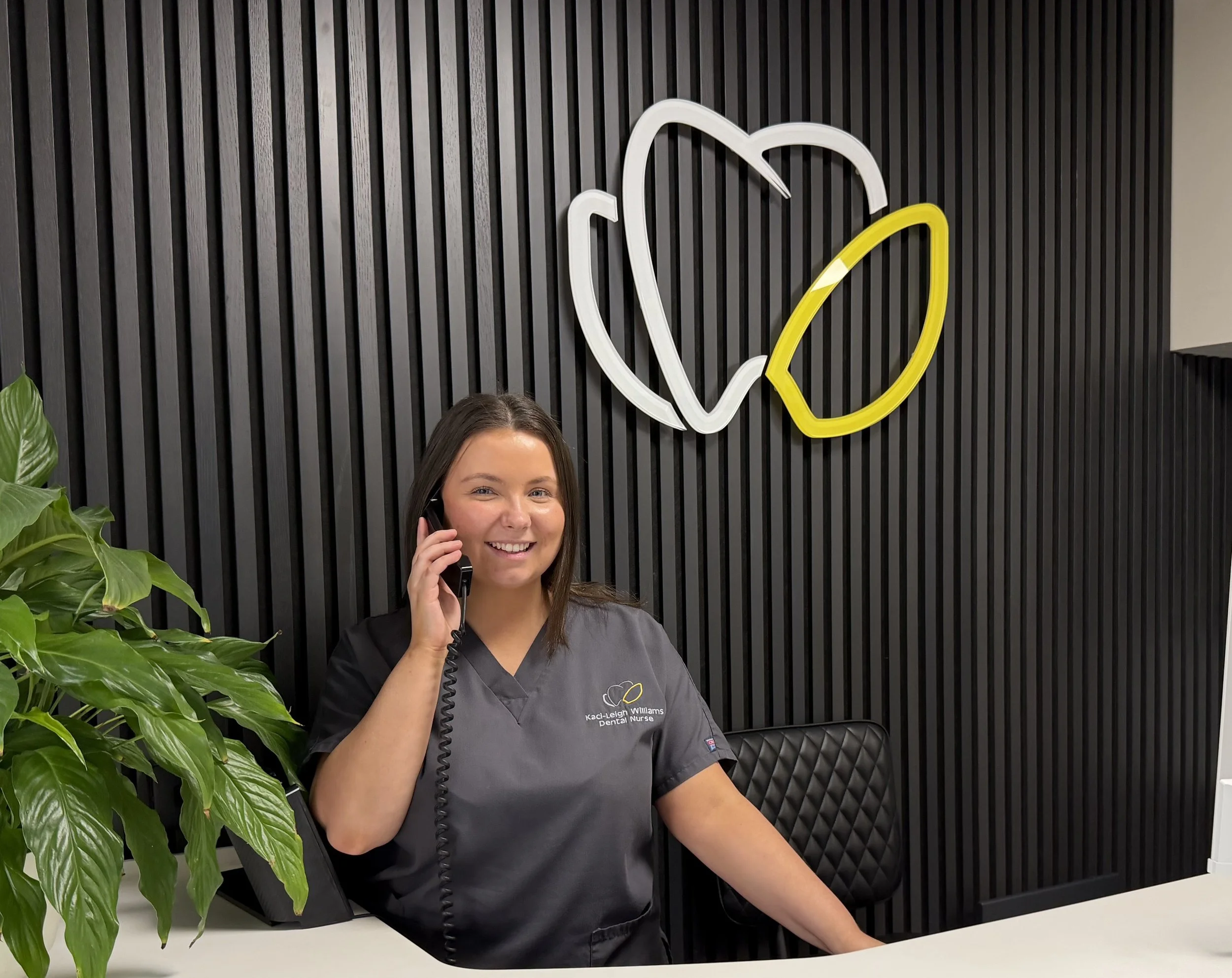 A smiling woman wearing dental scrubs sitting at a reception desk, talking on the phone in a dental office with a black slatted wall and neon fruit outline in the background.