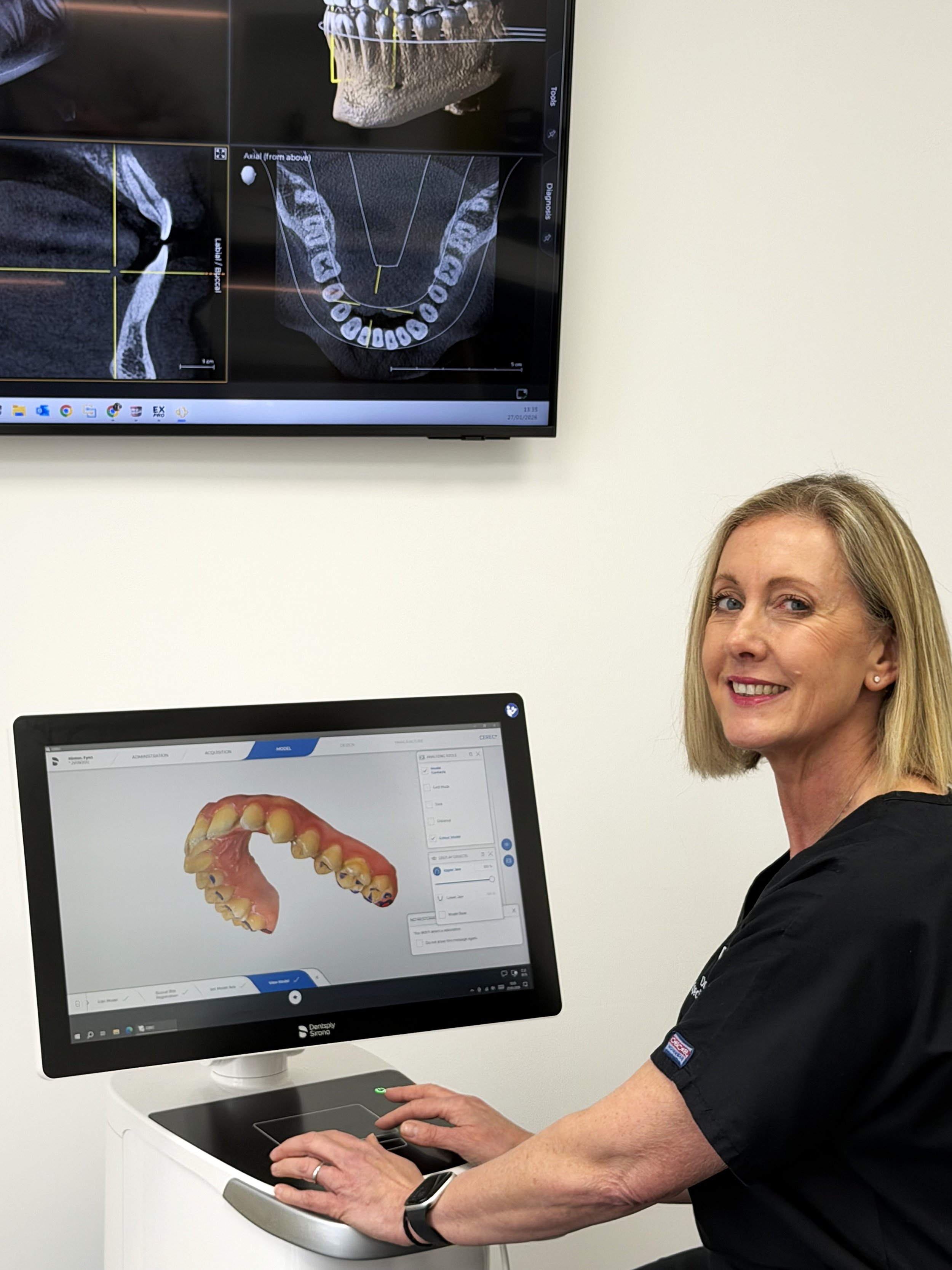 A woman sitting at a desk using a computer monitor, which displays a 3D digital model of a set of teeth with gum tissue in a dental software program, with a second screen in the background showing dental imaging scans.