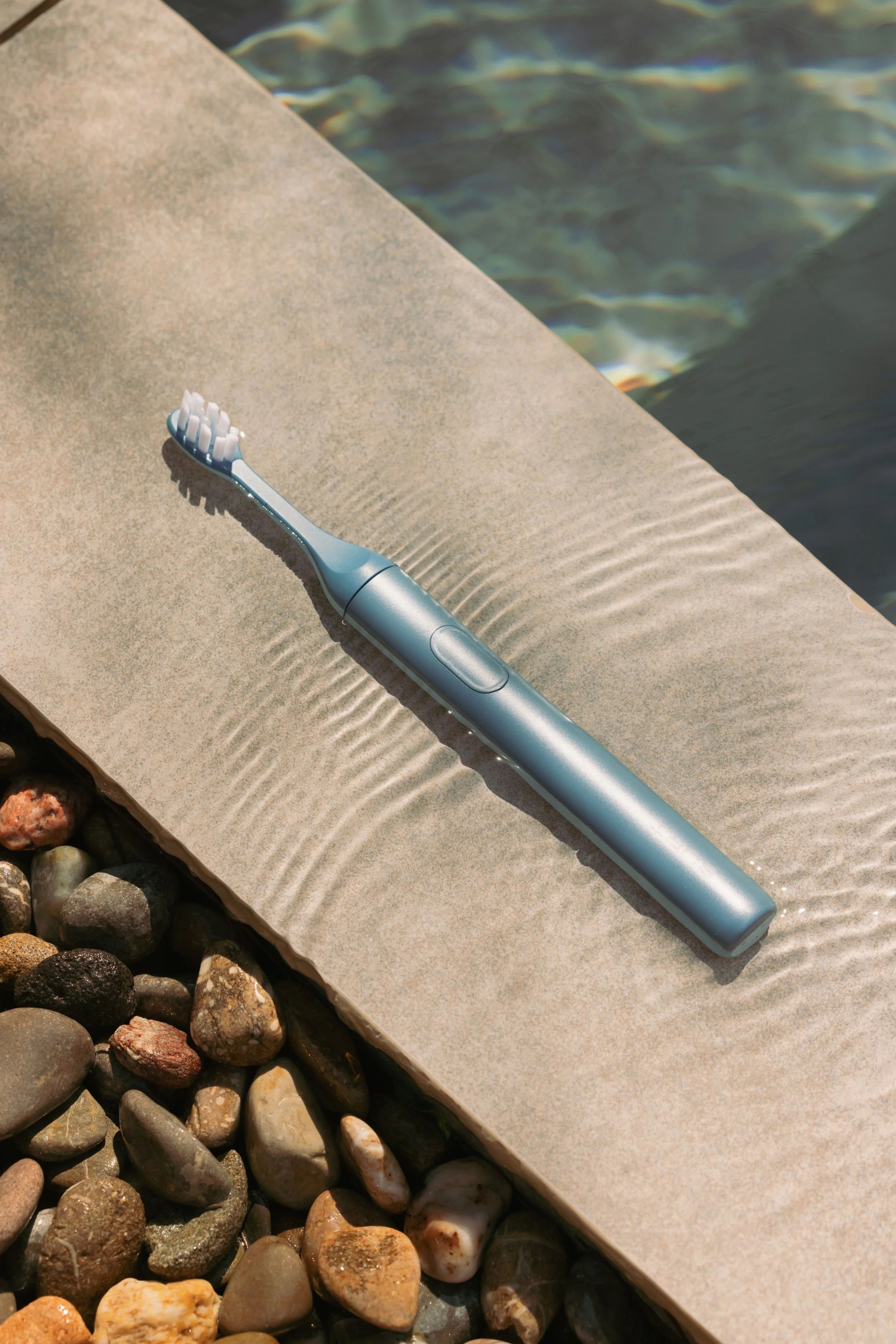 A light blue electric toothbrush resting on a concrete surface near a body of water, with pebbles visible in the corner.
