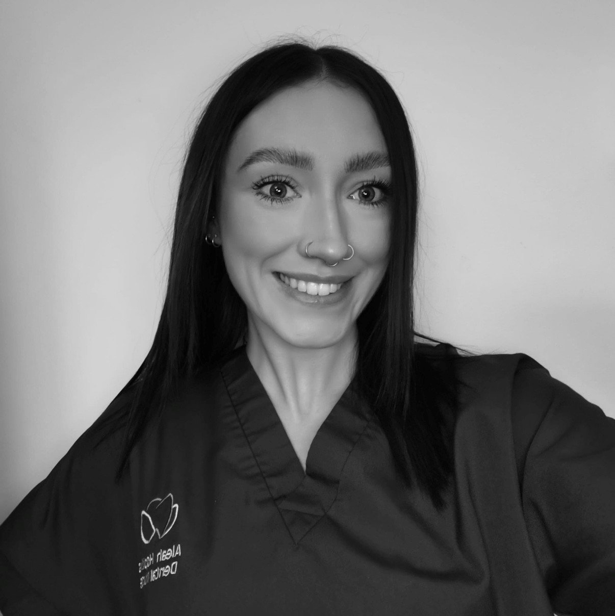 A woman with dark hair, wearing a dark uniform with an embroidered logo, smiling while taking a selfie against a plain wall.