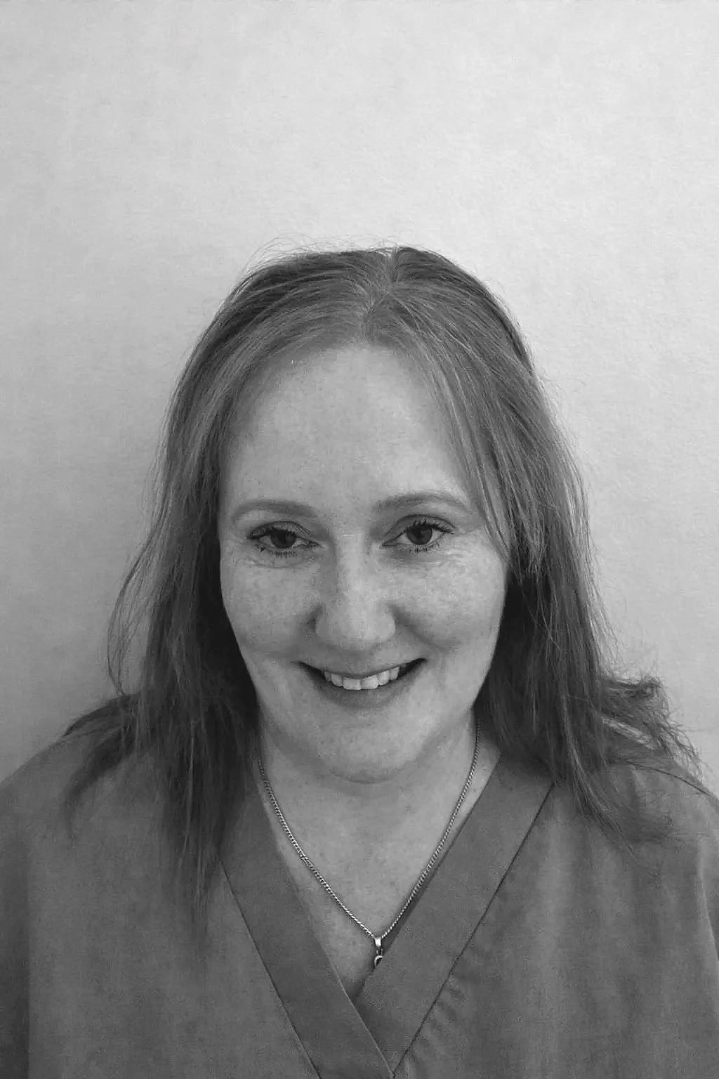 Black and white photo of a smiling woman with shoulder-length hair wearing a necklace, standing against a plain wall.
