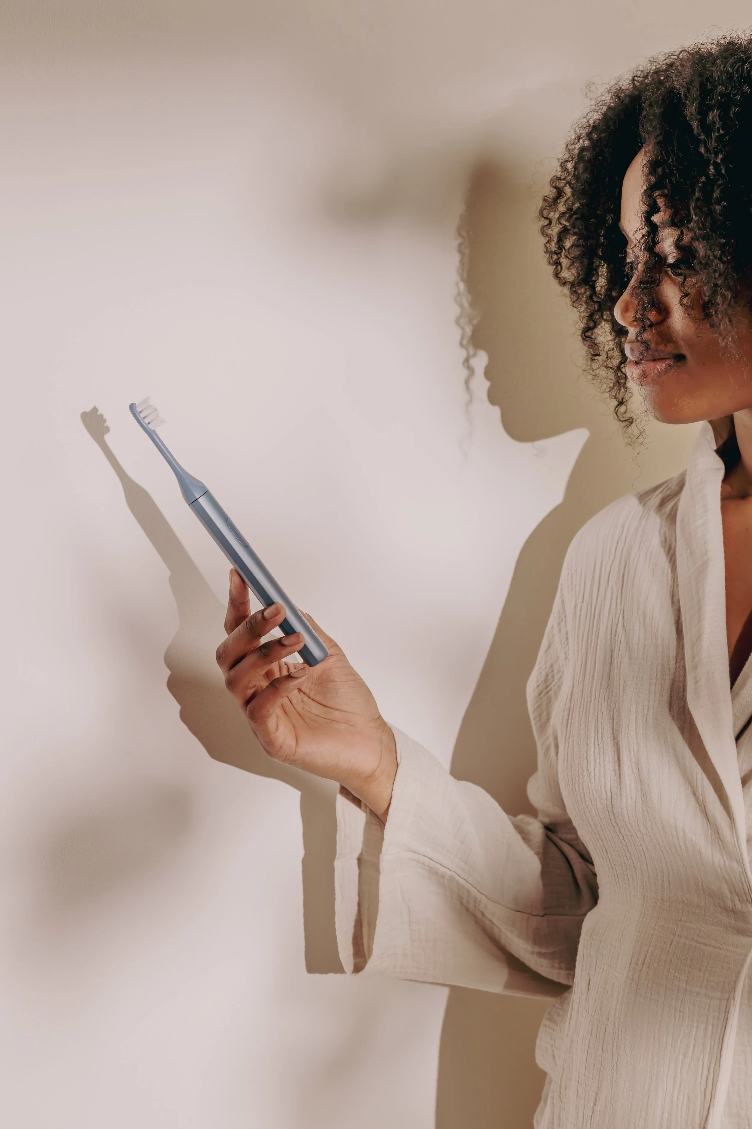 A woman with curly hair holding a blue electric toothbrush, standing against a beige wall.