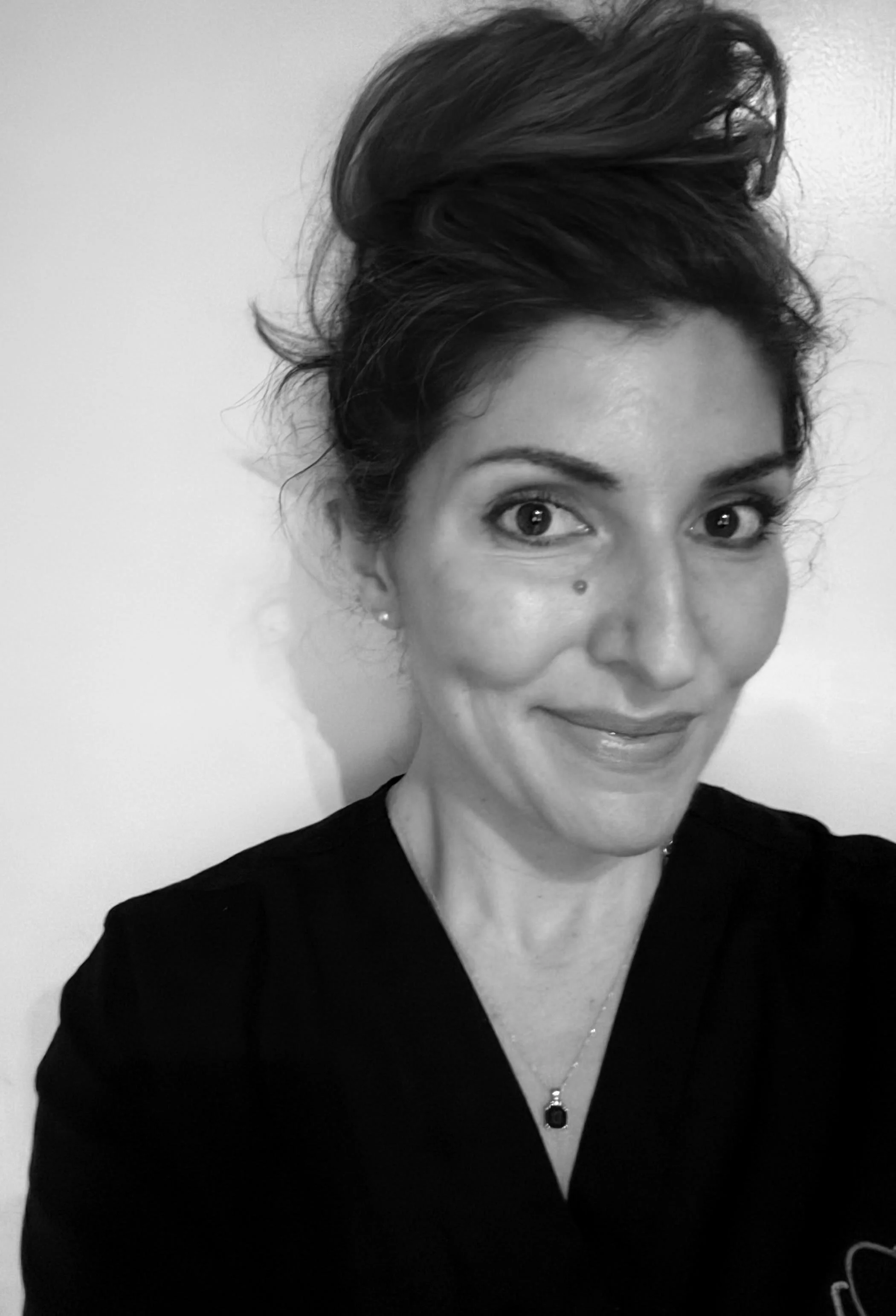 A woman with dark hair styled in an updo, smiling, wearing a black top and a necklace with a pendant, against a plain background.