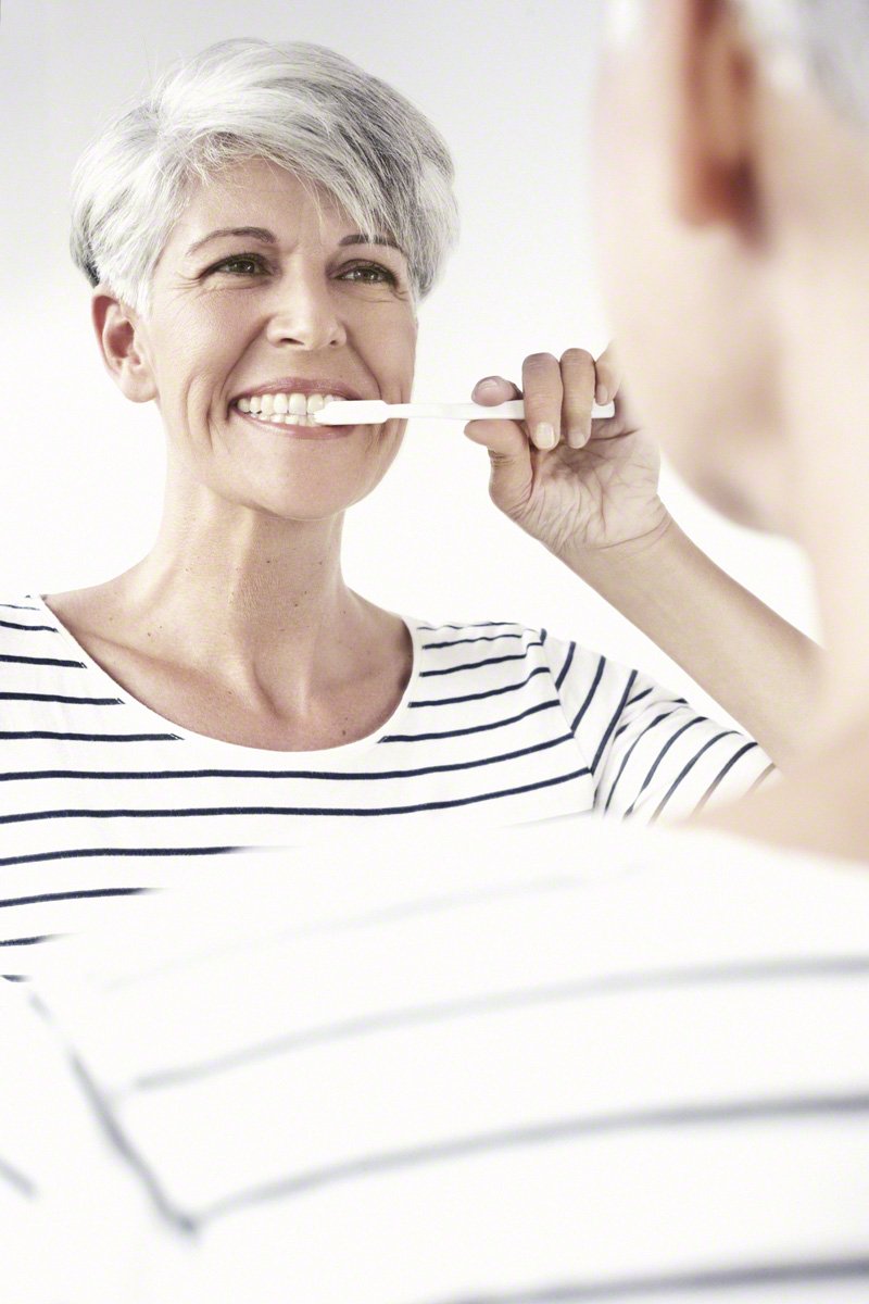 An older woman with gray hair smiling as she brushes her teeth in front of a mirror.