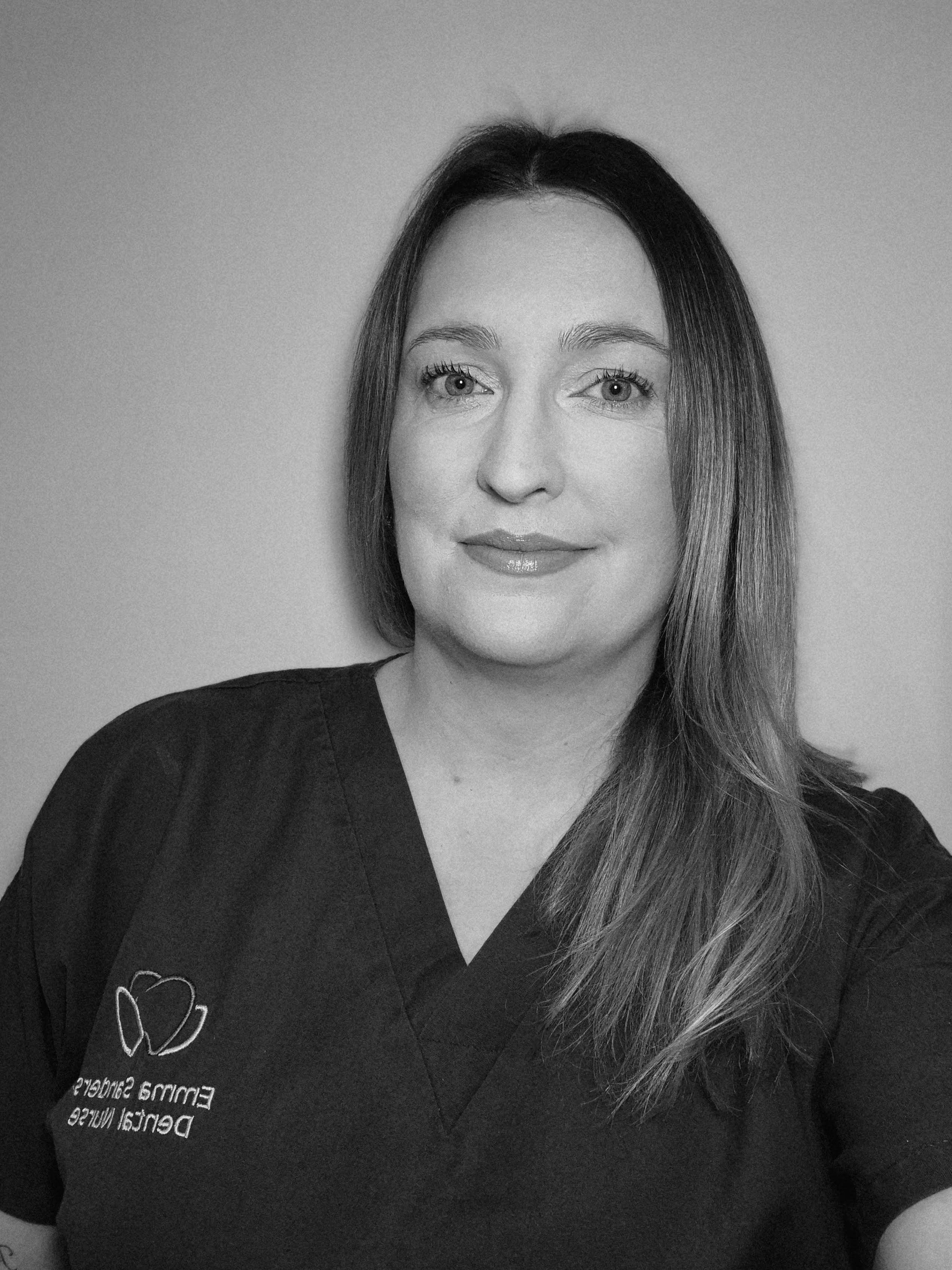 Black and white photo of a woman with straight hair wearing a dark medical scrub top with a logo on the chest.