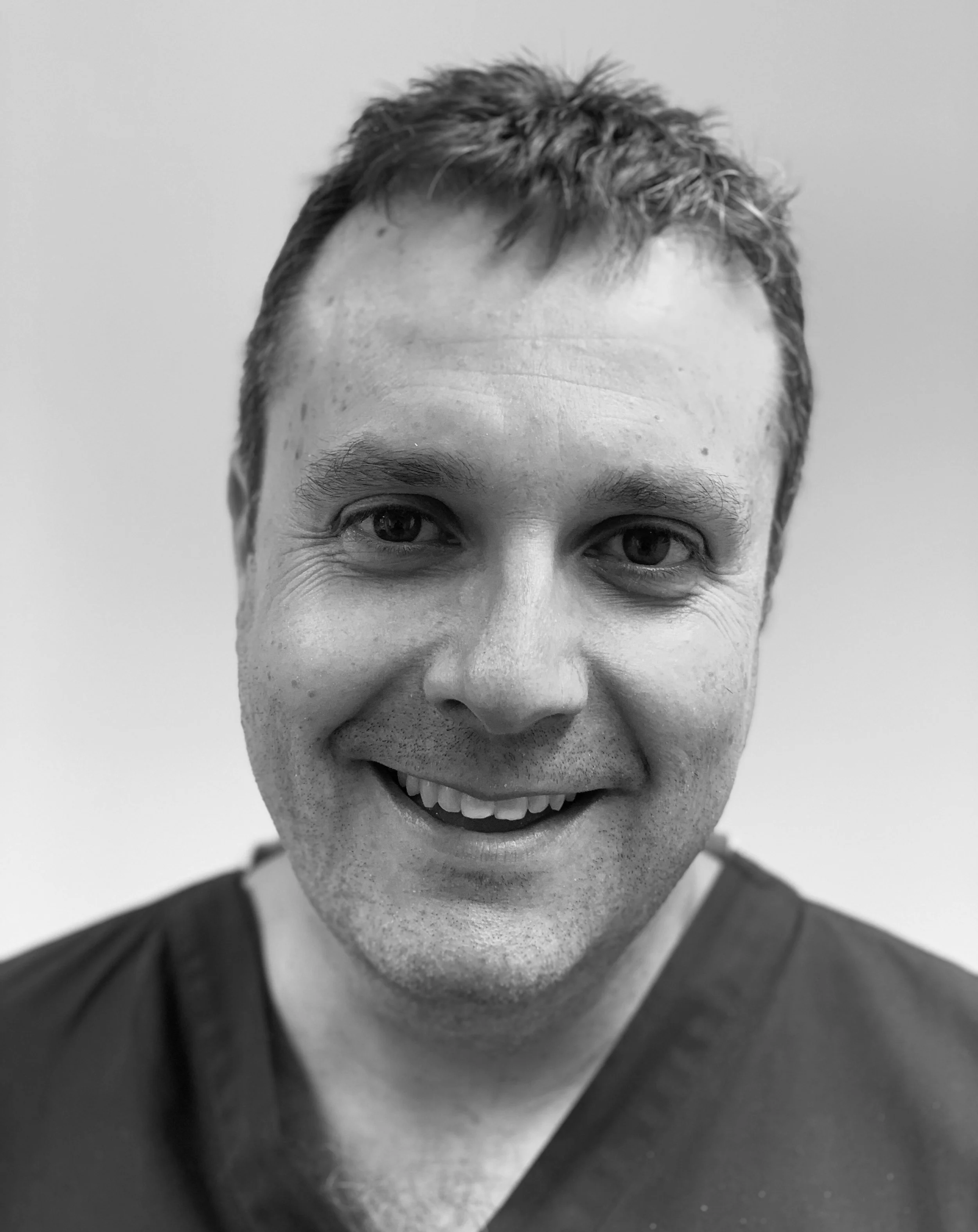 Black and white photo of a smiling man with short hair, wearing a dark shirt, looking directly at the camera.