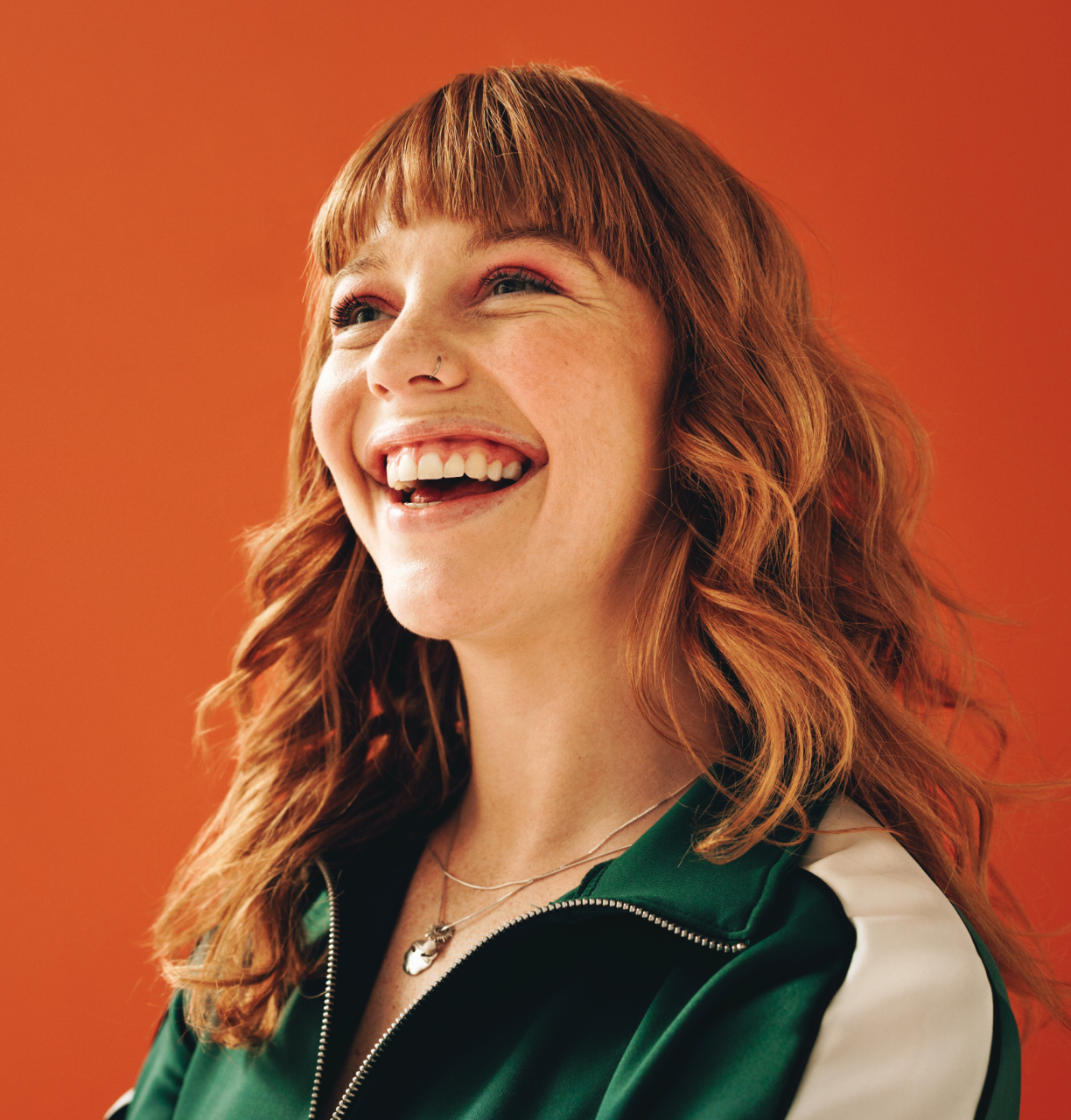 A young woman with red hair and a nose piercing smiling and laughing, wearing a green jacket with a white stripe on the sleeve, against an orange background.