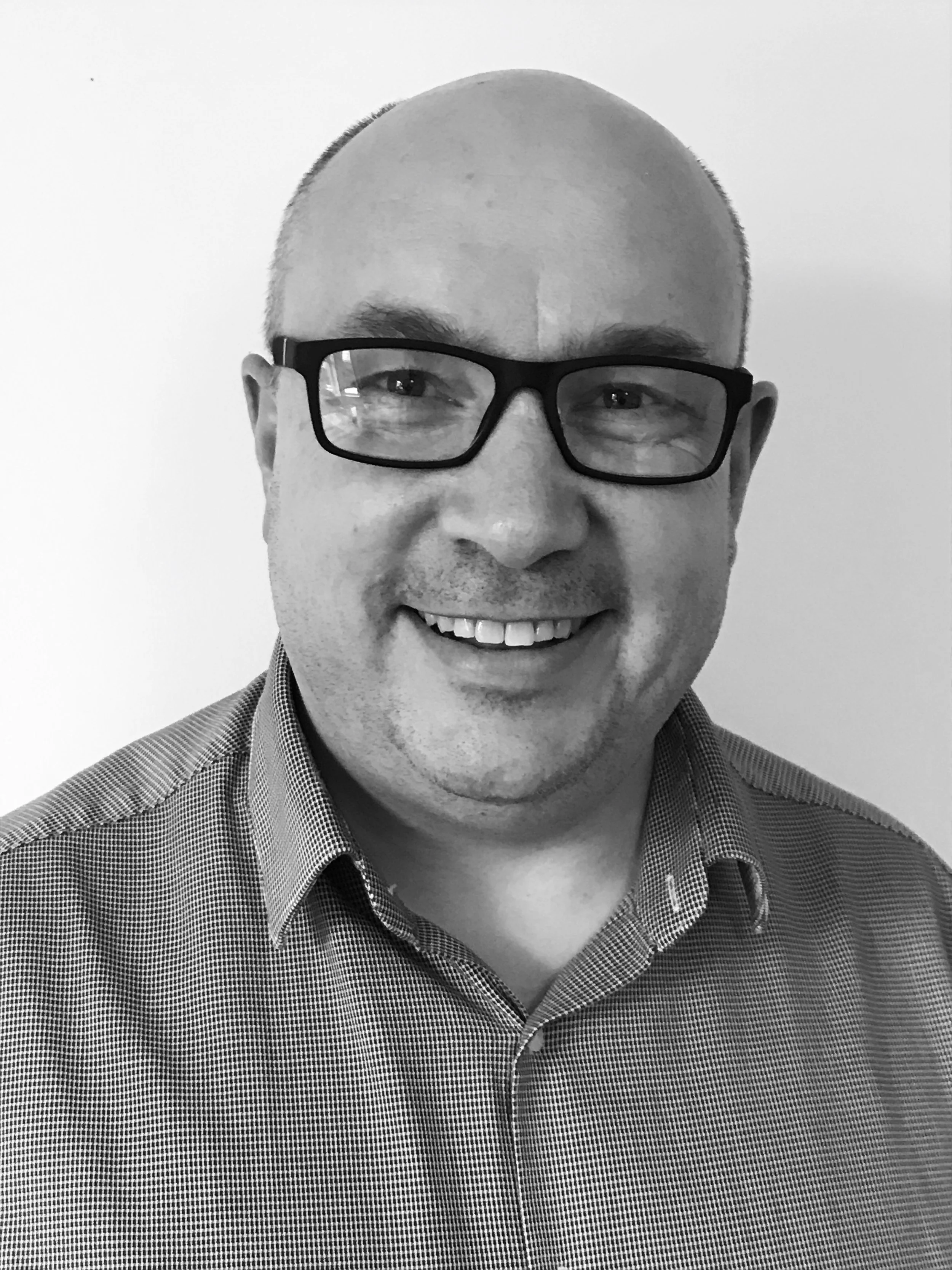 A smiling man with glasses and a checkered shirt, facing the camera, black and white