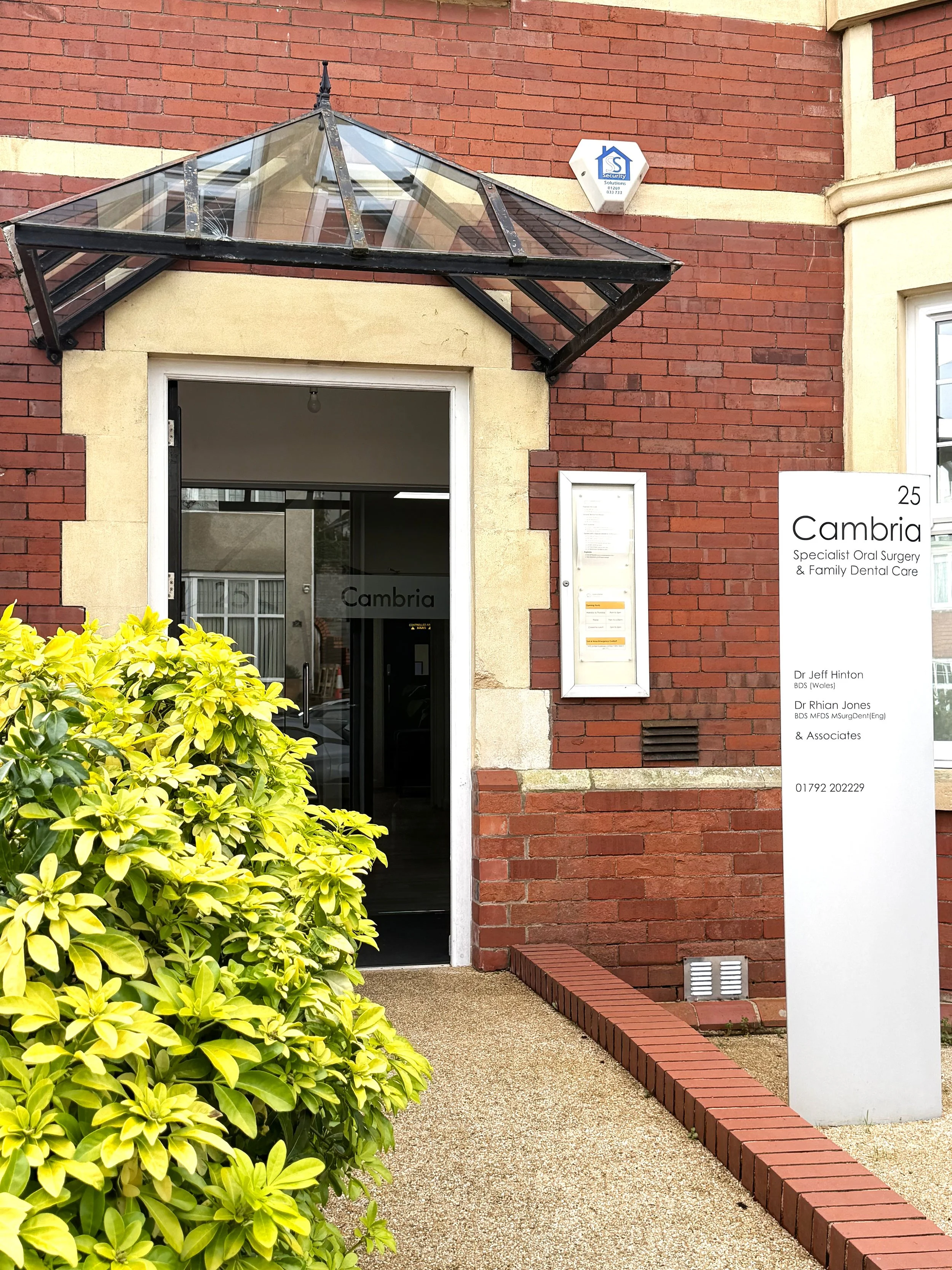 Entrance to Cambria dental clinic with glass canopy, sunny yellow bush, white sign with black text, brick and stone building.