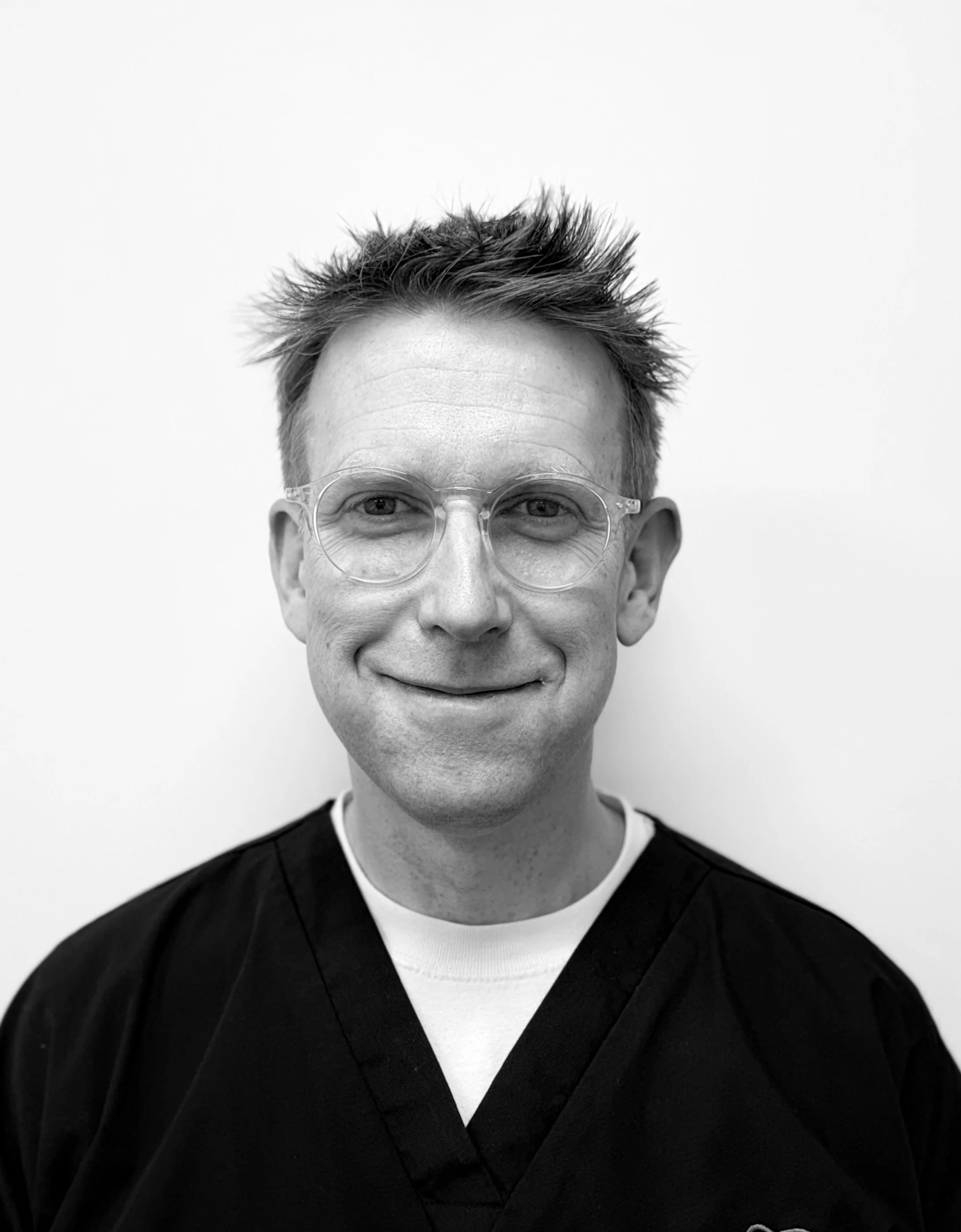 Black and white portrait of a man smiling, wearing glasses and a dark medical scrub top, standing against a plain white background.