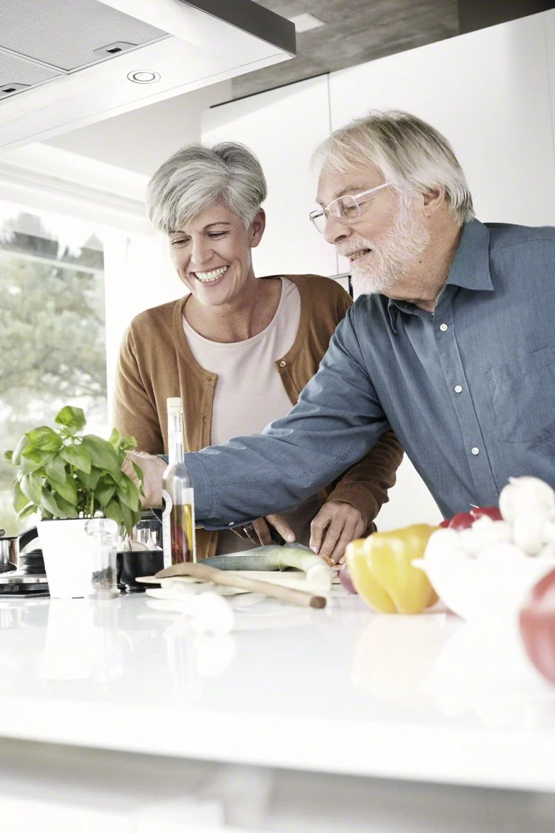 An older couple cooking together in a bright, modern kitchen, smiling as they prepare food.