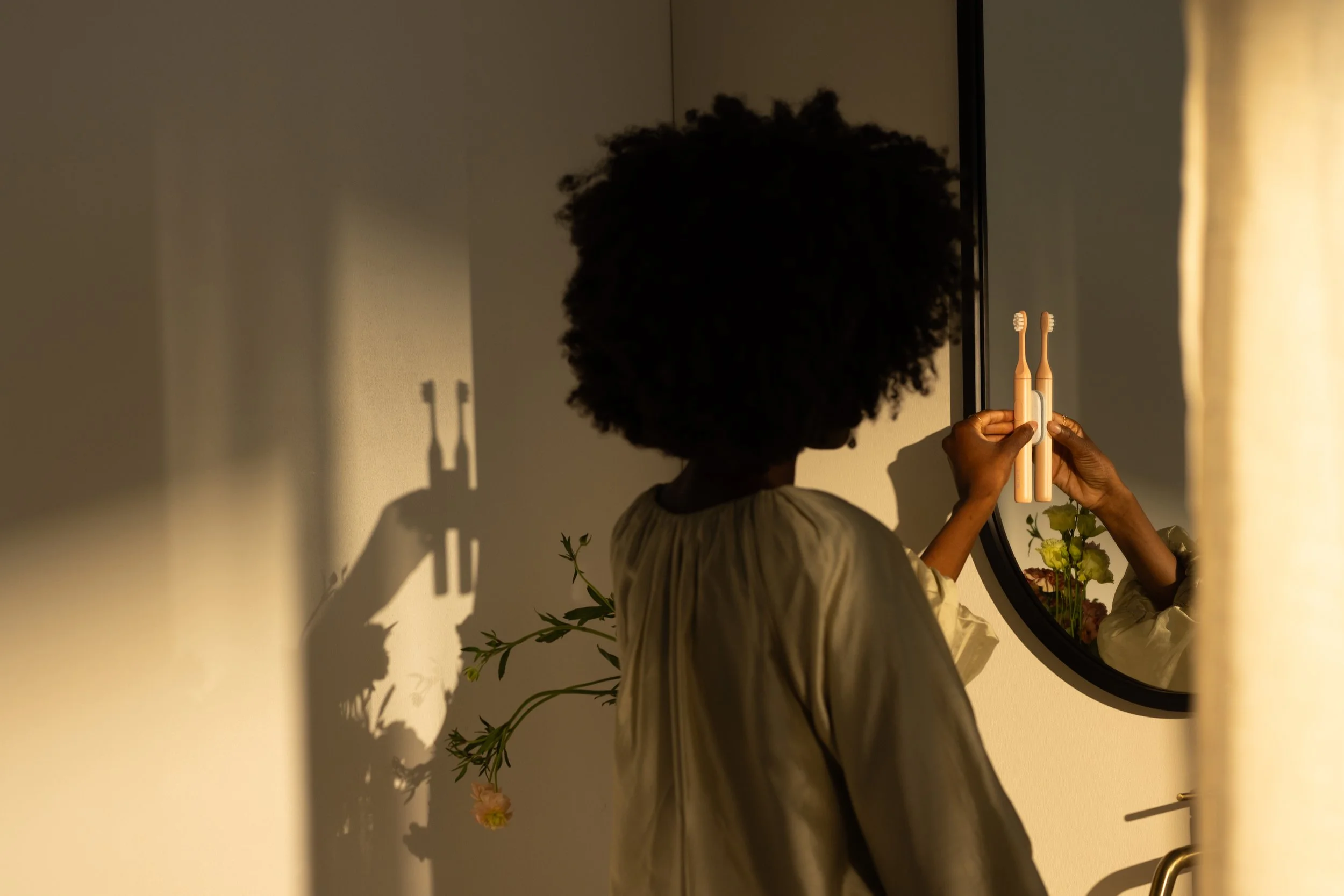 A woman with dark curly hair in a cream-colored shirt holds two toothbrushes in front of a round mirror in a bathroom. Her reflection is visible, and a plant with green leaves and pink flowers is on the wall behind her. The scene is softly lit, creating shadows on the wall.
