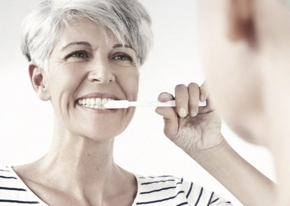 A woman with short gray hair brushing her teeth while smiling.