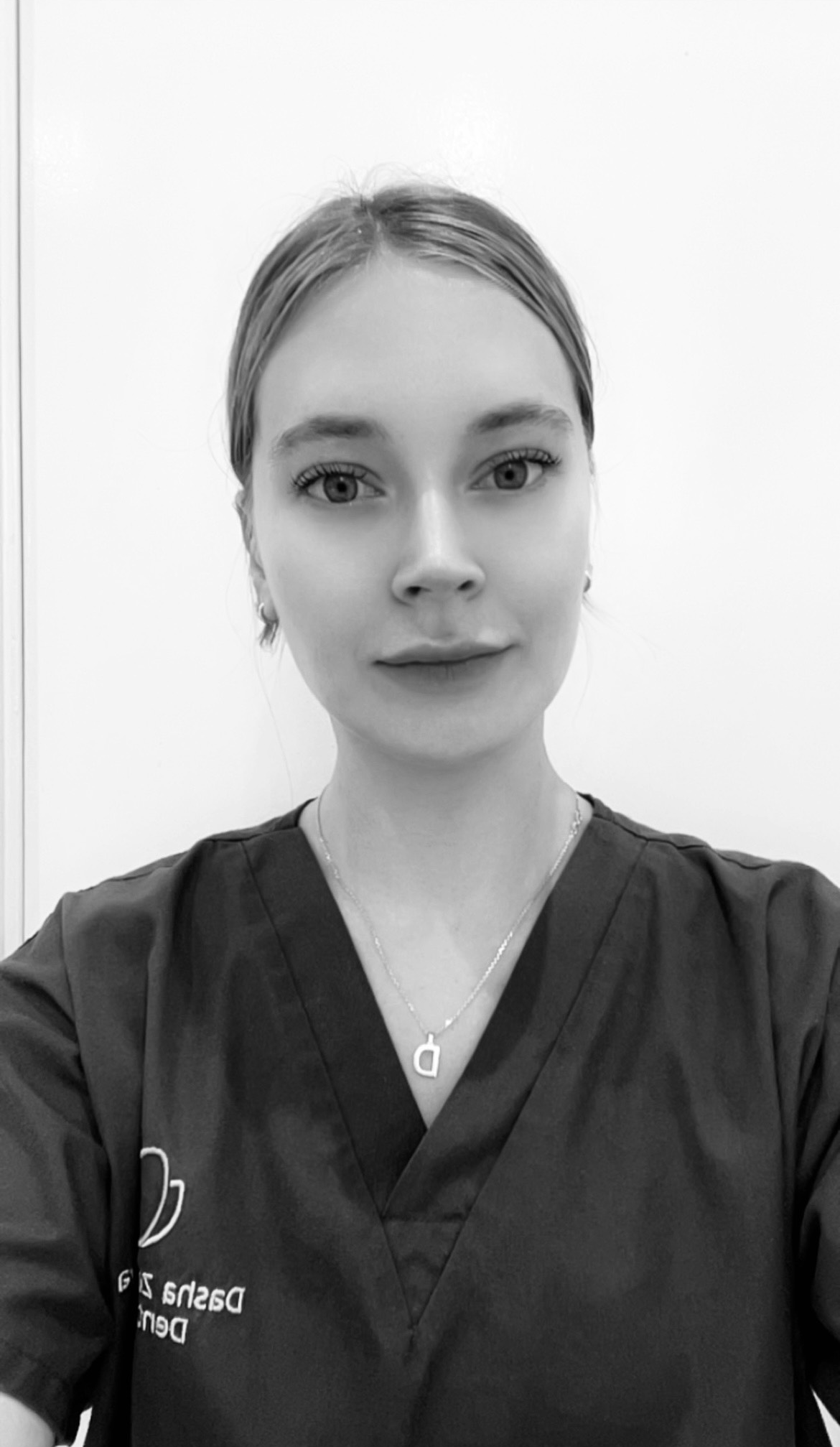 Black and white photo of a young woman with light-colored hair pulled back, wearing a dark medical scrub top with a logo and name embroidered on it and a necklace with a key pendant shaped like the letter 'D'.