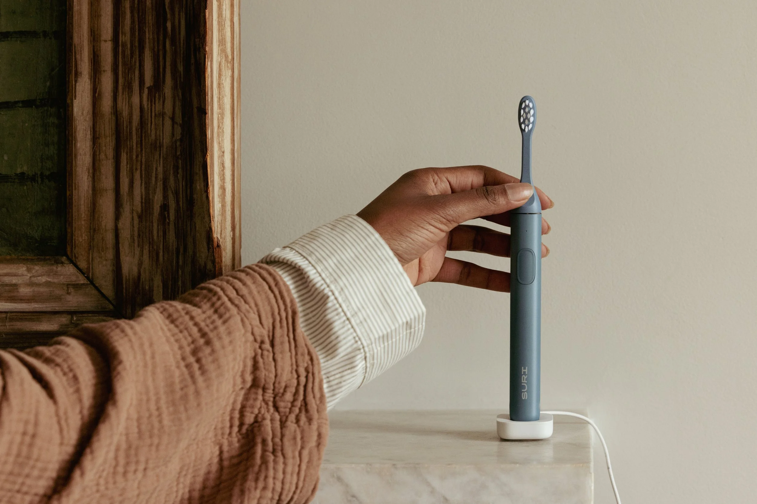A person holding a blue electric toothbrush beside a charging base on a marble surface, with a beige wall in the background.