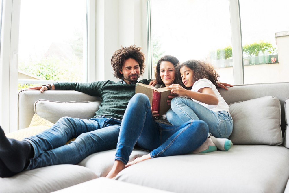 Three people sitting on a sofa near a large window, reading a book together.