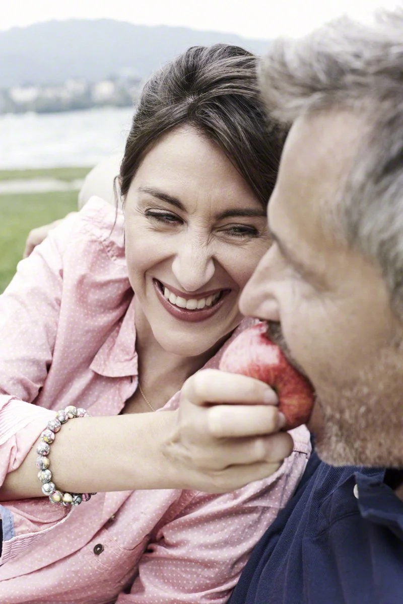 A woman with brown hair smiling and holding a red apple close to a man's mouth, outdoors with a grassy field and water in the background.