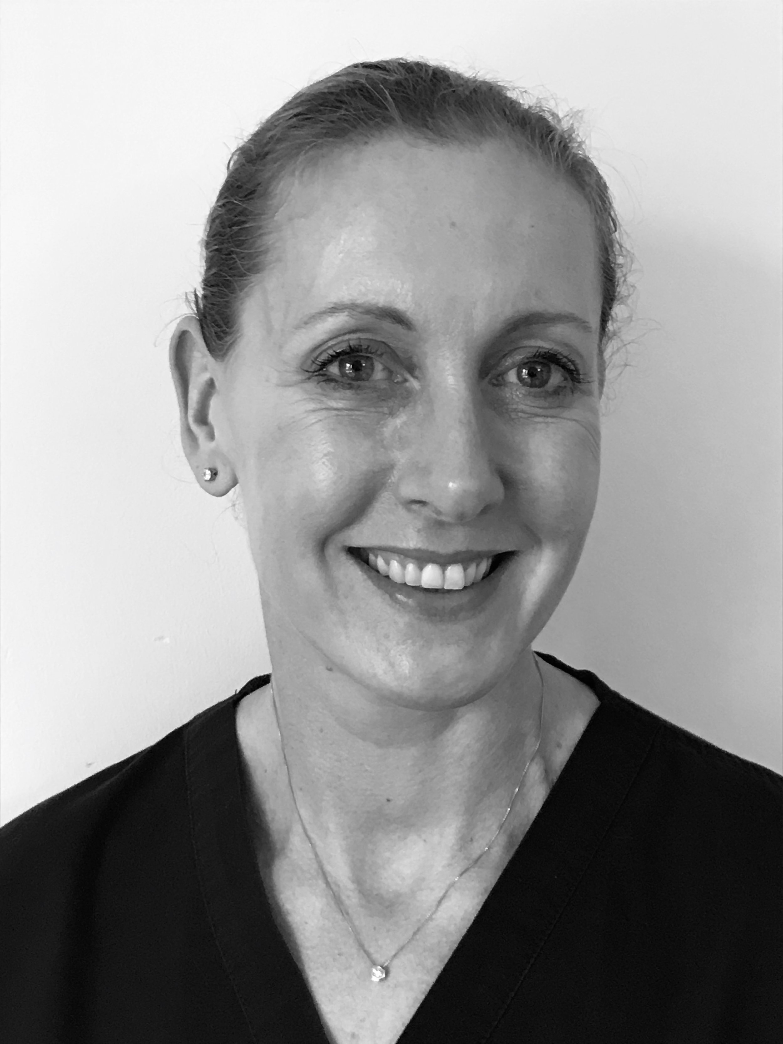 Black and white portrait of Rhian Jones with short hair, wearing a dark top, small earrings, and a necklace, against a plain background.
