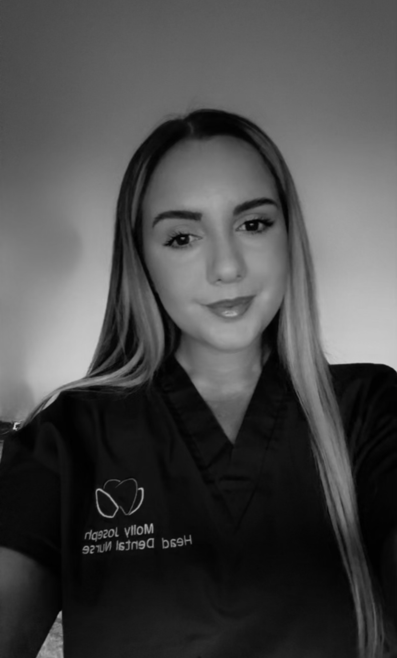 Black and white photo of a woman with long hair, wearing a uniform with a logo and the text 'Molly Joseph Head Dental Nurse' on it, smiling at the camera.