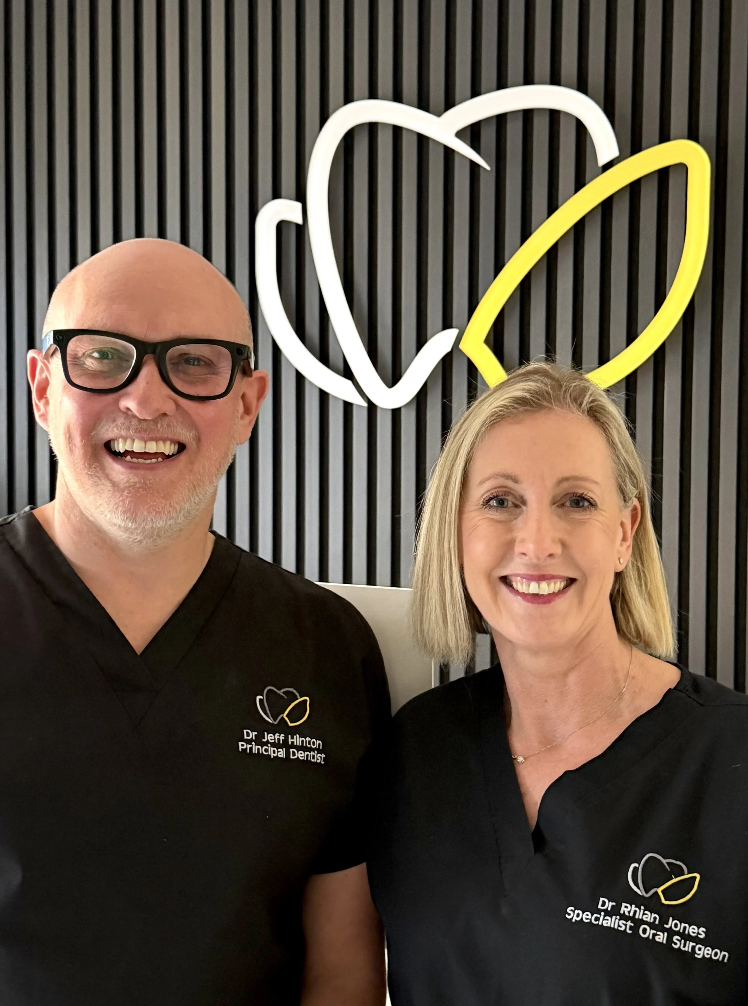 Two smiling dental professionals, a man with glasses and a woman, stand in front of a black wall with a neon sign of a tooth and a crown.