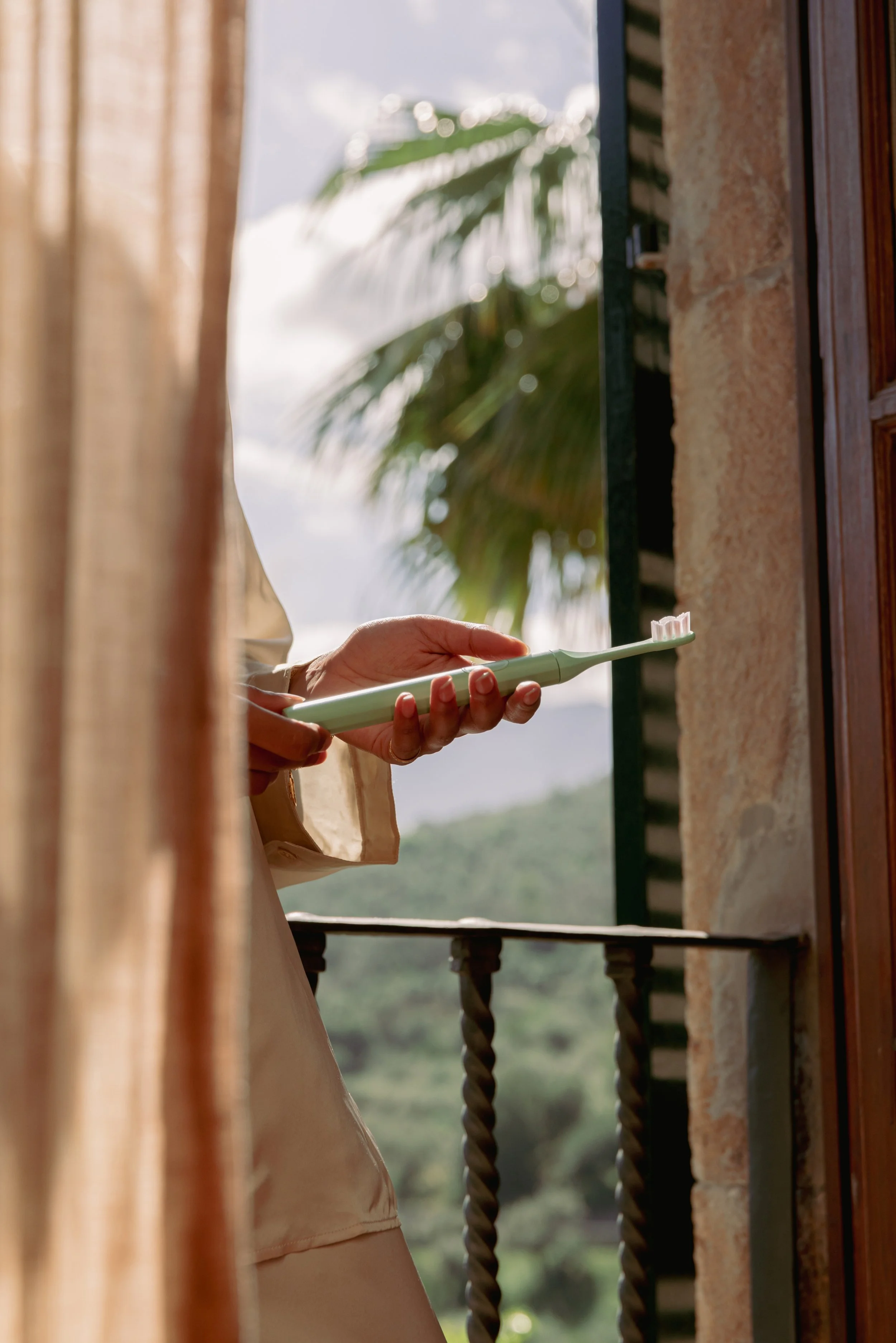 Person holding a green toothbrush on a balcony with a tropical background.