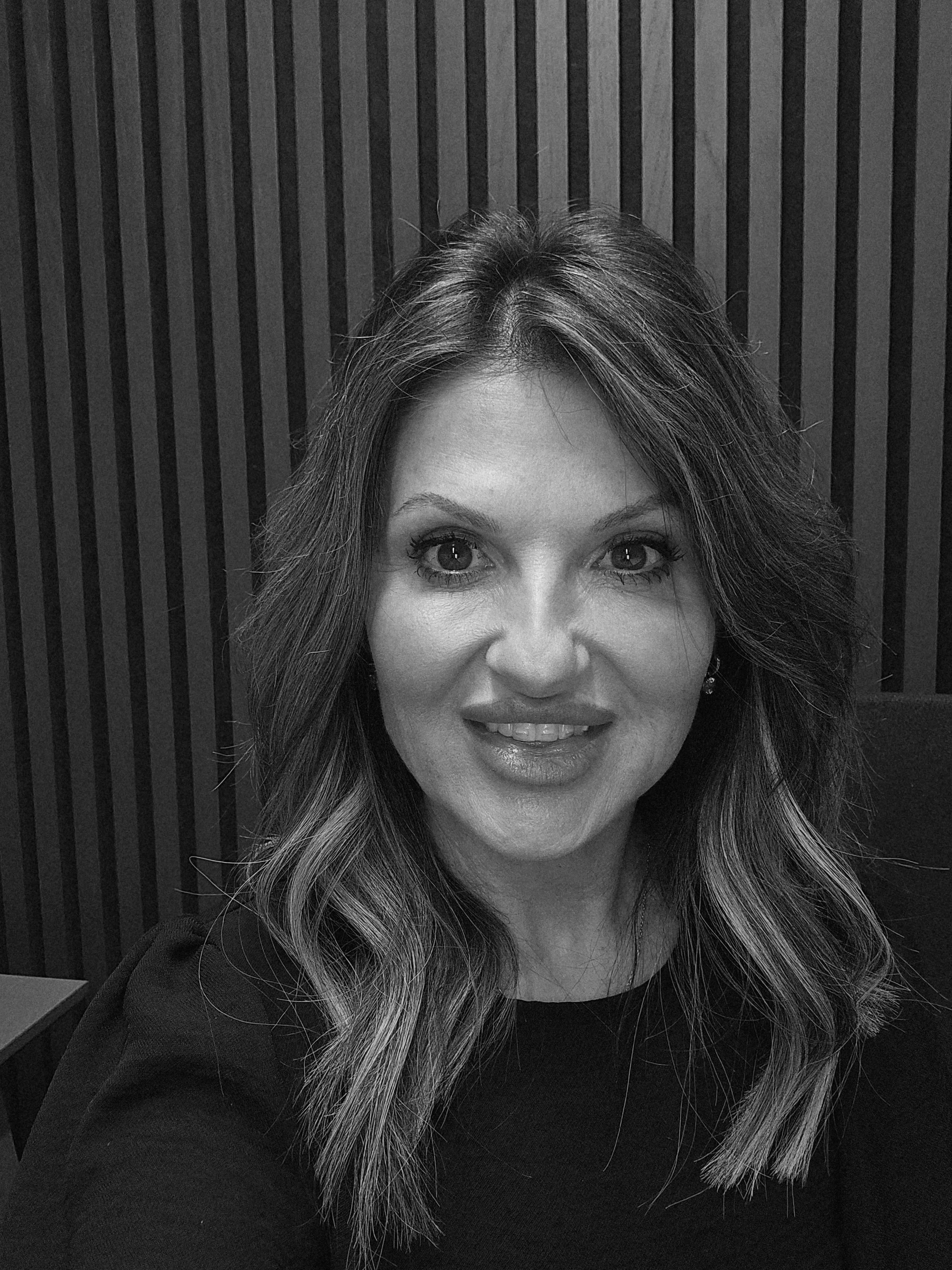 A woman with wavy hair and makeup smiling for a selfie in front of a striped wall in black and white.