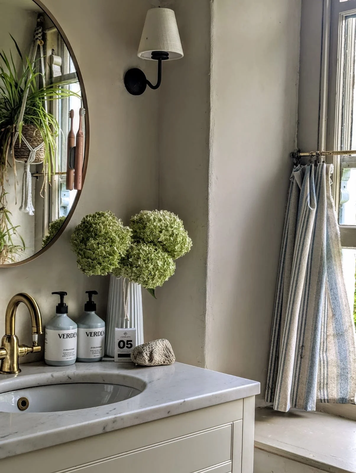 A bathroom sink area with a marble countertop, two bottles of hand soap, a vase with green hydrangea flowers, a soap sponge, a large mirror, a wall-mounted light fixture with a white lampshade, and a window with striped curtains.