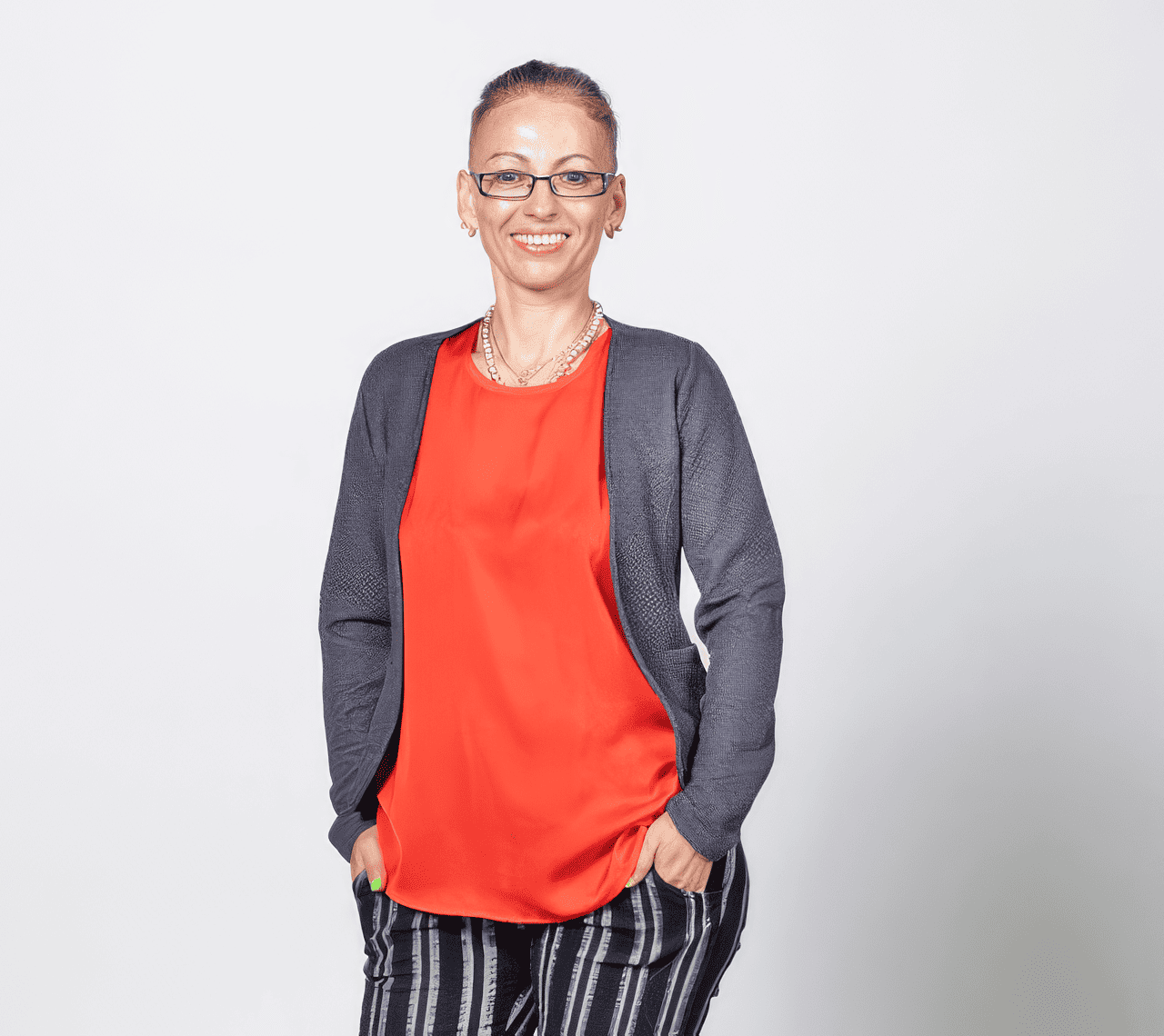 A smiling woman with glasses, wearing a red blouse, gray blazer, and striped pants, standing against a plain white background.