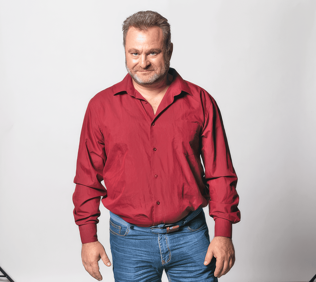 A middle-aged man with gray hair and beard wearing a red button-down shirt and blue jeans, standing against a plain white background.