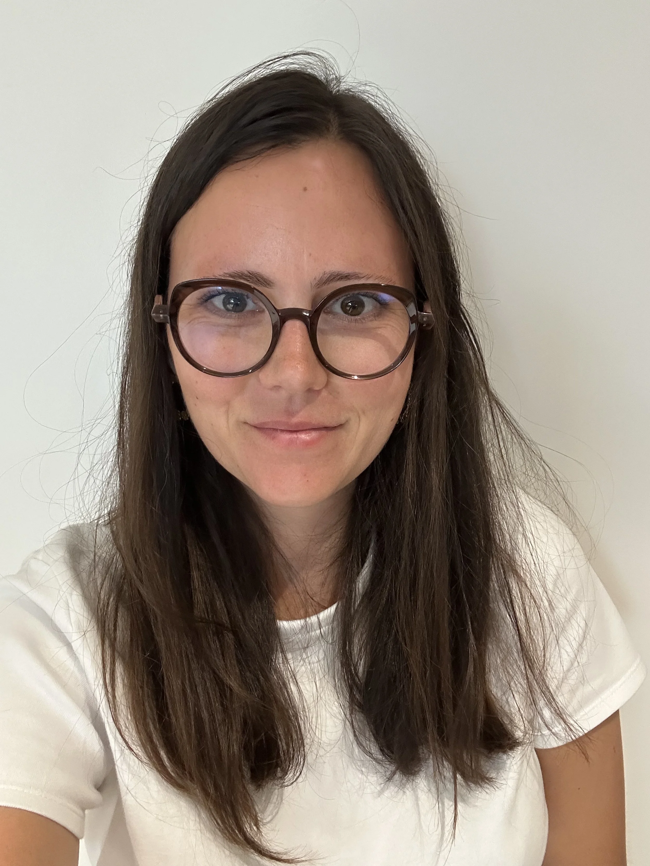 Une jeune femme portant des lunettes rondes, avec de longs cheveux bruns, souriant, portant un t-shirt blanc, en arrière-plan un mur blanc.