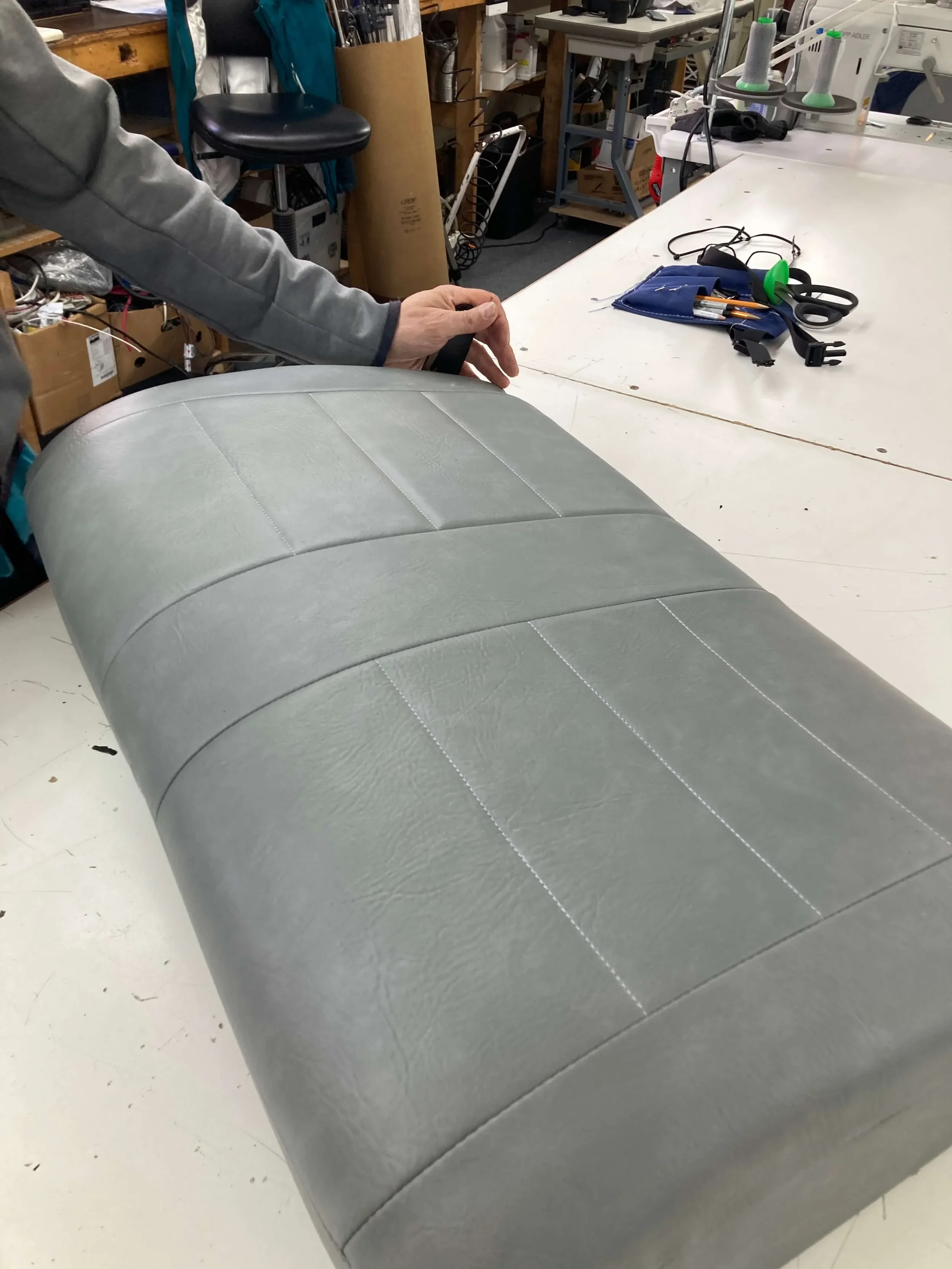Person working on reupholstering or inspecting a light gray, vinyl or leather car bench seat in a workshop with tools and supplies on the work surface.