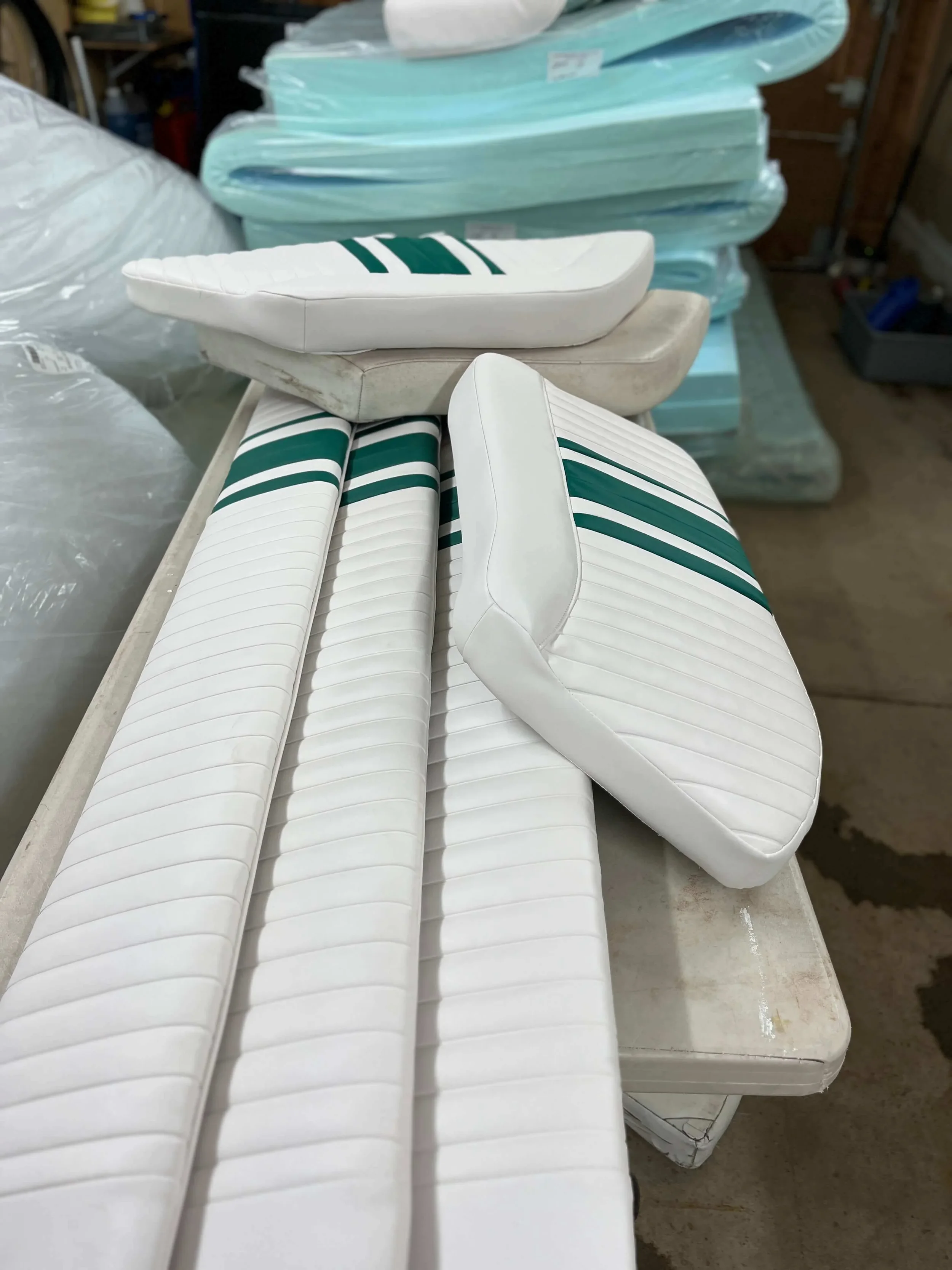 Multiple white chair cushions with green stripes and some plain white cushions stacked on a table in a storage or workshop area.