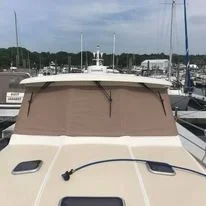 View of the bow of a boat with a Robie Canvas custom windshield cover done in beige over the front area, docked at a marina with other boats and trees in the background.