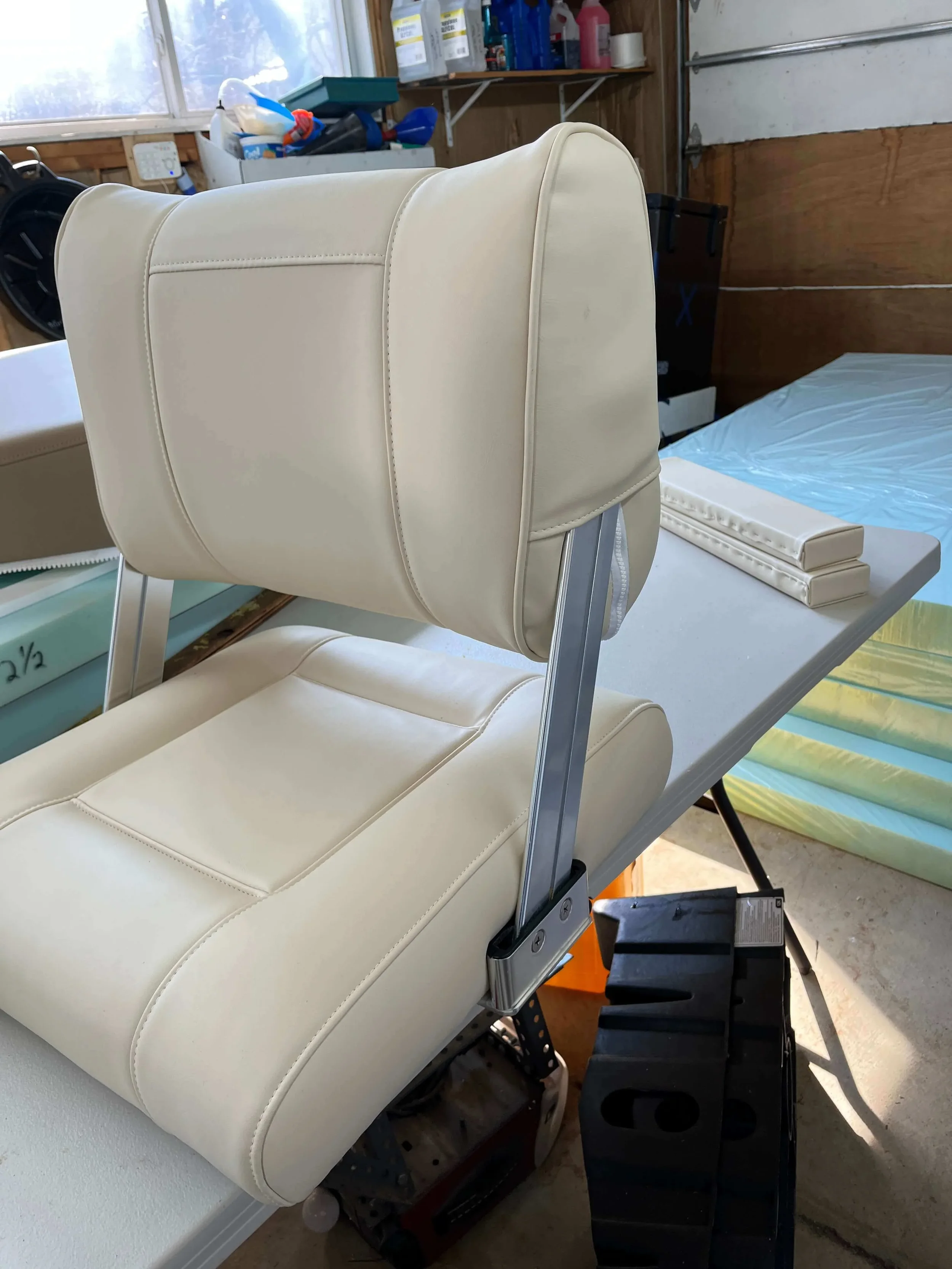 A cream-colored chair with a padded backrest and seat, mounted on a metal frame, positioned in a workshop or garage setting with tools and other items in the background.