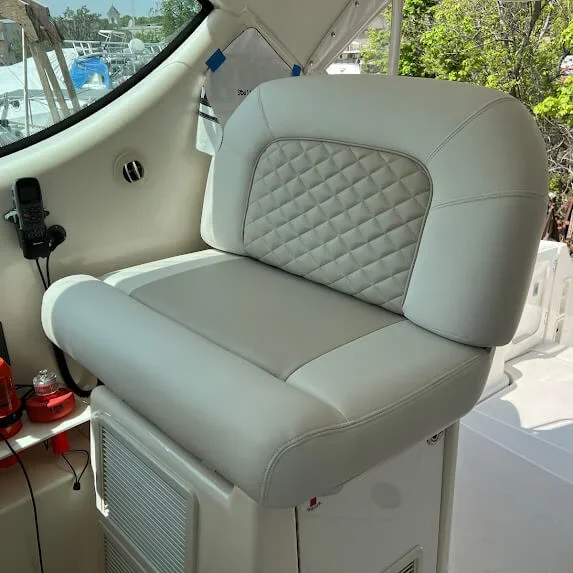 A white custom fabricated Robie Canvas cushioned seat with diamond-stitched gray backrest is inside a boat, near a window showing a marina with boats and trees outside.