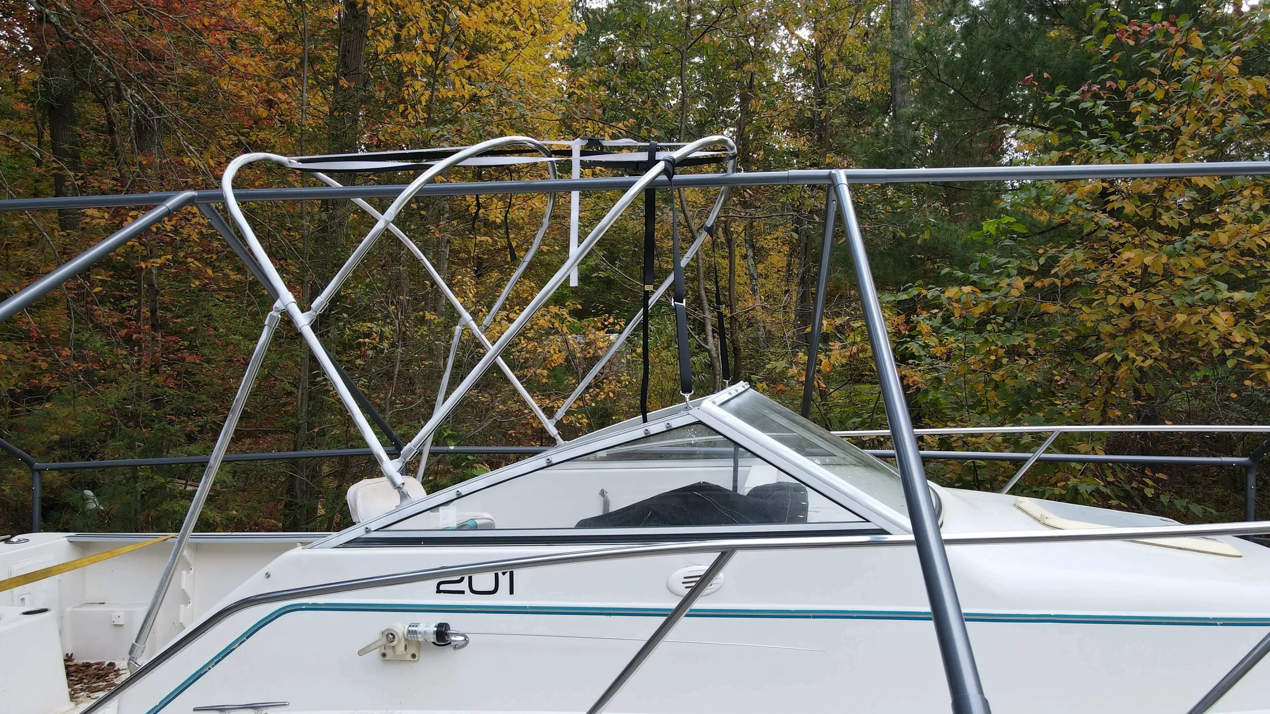 A white boat with a Robie Canvas custom bent stainless bimini frame structure on top, parked near trees with autumn-colored leaves.