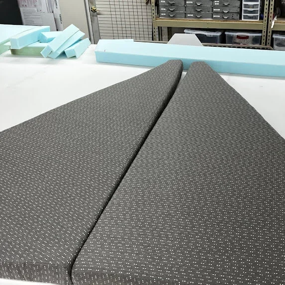 Two gray upholstered bow cushions with small white polka dots on a white table, surrounded by blue foam pieces in the Robie Canvas workshop ready for installation.