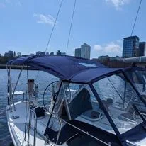 A boat docked at a marina on a clear day with blue skies and city buildings in the background, highlighting the new custom dodger, connector flap, and rear 3 bow bimini custom fabricated by Robie Canvas.