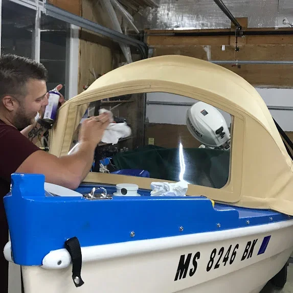 A Robie Canvas employee working on a small boat with a tan dodger canopy inside a garage, with various tools and equipment around.