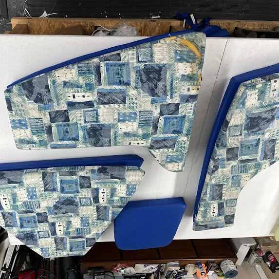 Parts of a boat interior with patterned upholstery and blue trim, including a seat cushion and a panel, on a white surface in a workshop.