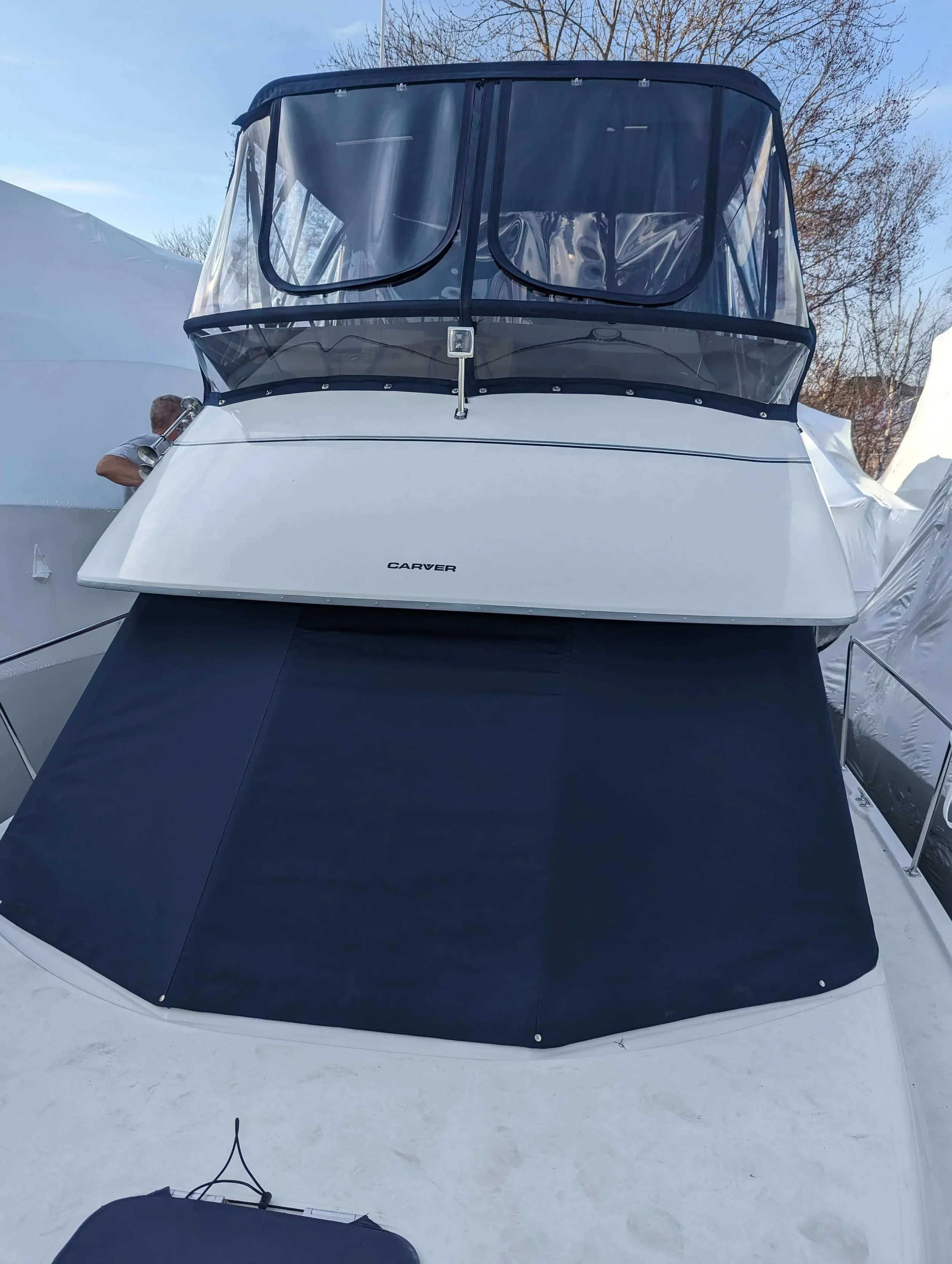 Front view of a white and black boat with a Robie Canvas custom windshield cover done in black mesh, docked at a marina. The brand name 'Carver' is visible on the front of the boat. A person is partially visible on the left side of the image.
