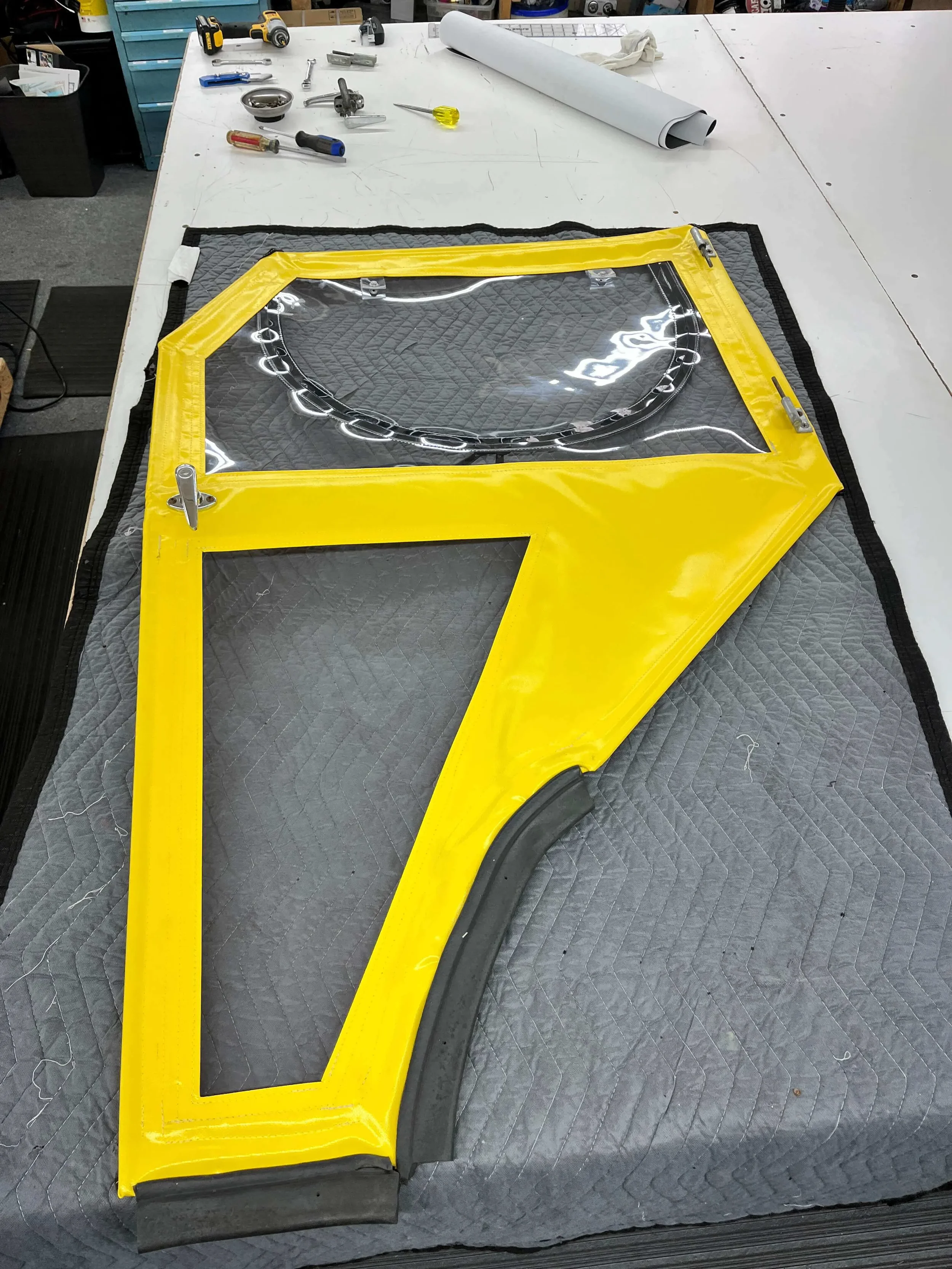A yellow fabric tractor door panel, repaired by Robie Canvas, secured with metal clips on a padded work surface. Tools and materials are visible on the nearby white worktable.