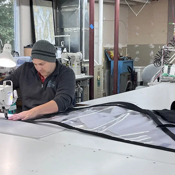 A man sewing what appears to be a large inflatable or plastic item in a workshop or manufacturing space.