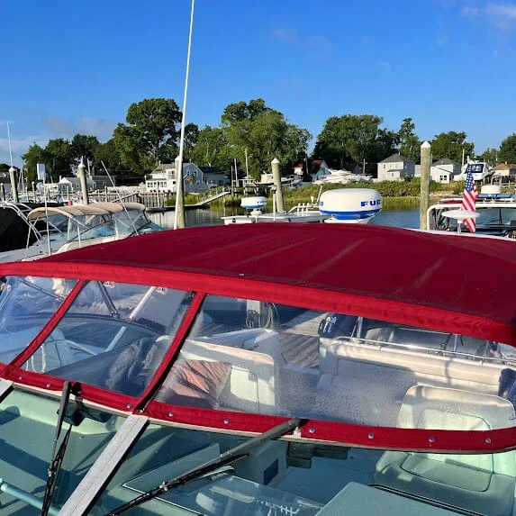 View of a boat with a red canopy docked at a marina with other boats and houses in the background, under a blue sky.
